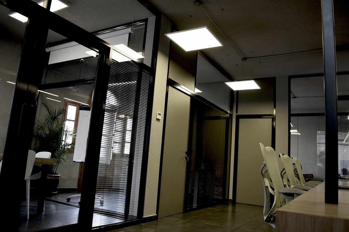 Modern office space with glass partition walls, ceiling lights, and chairs along a desk area.
