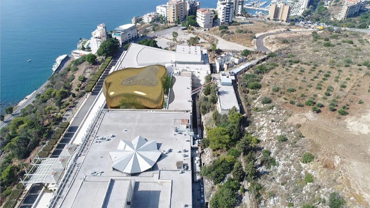 Aerial view of a city with a coastline and several buildings, including a distinctive golden, irregularly shaped structure on a rooftop, surrounded by trees and open land.