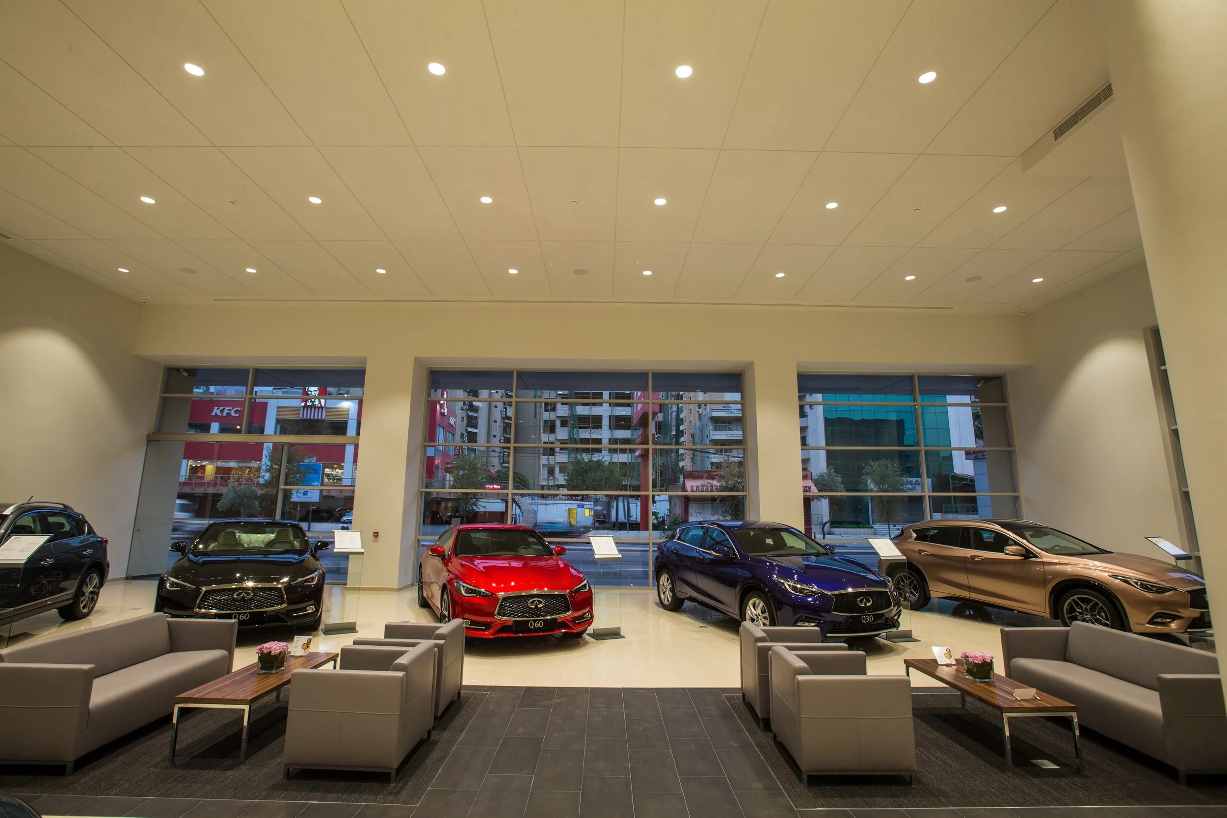 Interior view of a car showroom with several luxury cars displayed near large windows, seating area in the foreground, and city buildings visible outside.