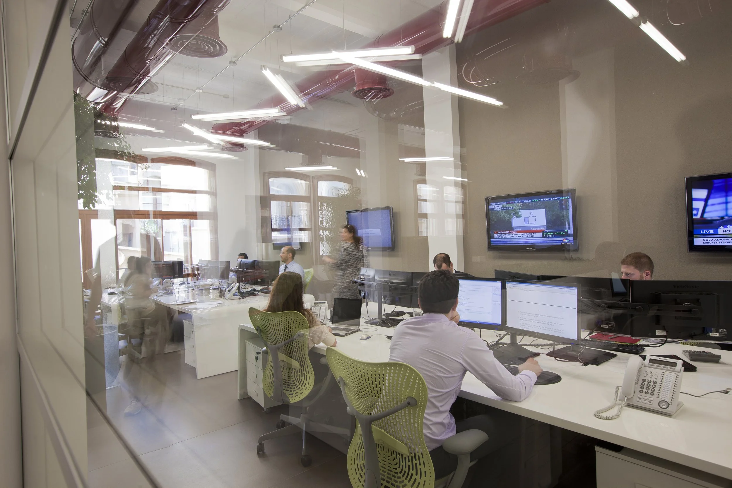 A modern office with multiple employees working at desks, some looking at computer screens, and a woman walking through the room. Large windows allow in natural light. Several television screens are mounted on the wall displaying news and stock infor