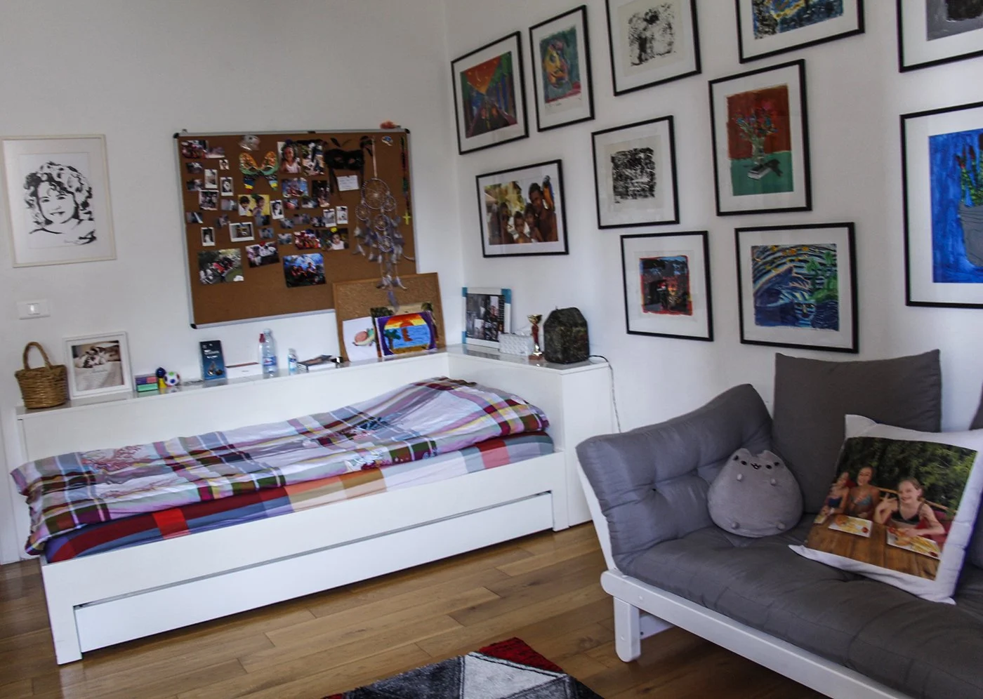 A bedroom with a single bed covered in a colorful plaid blanket, a gray futon with a pillow featuring a photo of two children, a corkboard with photos and decorations, a white shelf with framed pictures and bottles, and a wall decorated with multiple
