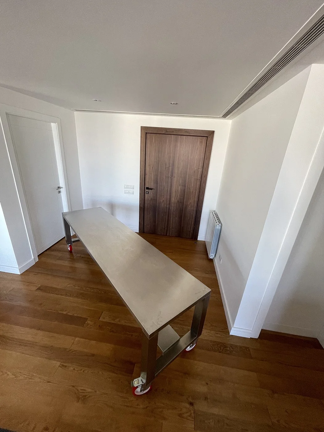 Interior of a room with wooden flooring, a wooden door, a white door, a radiator, and a long metal table with wheels.