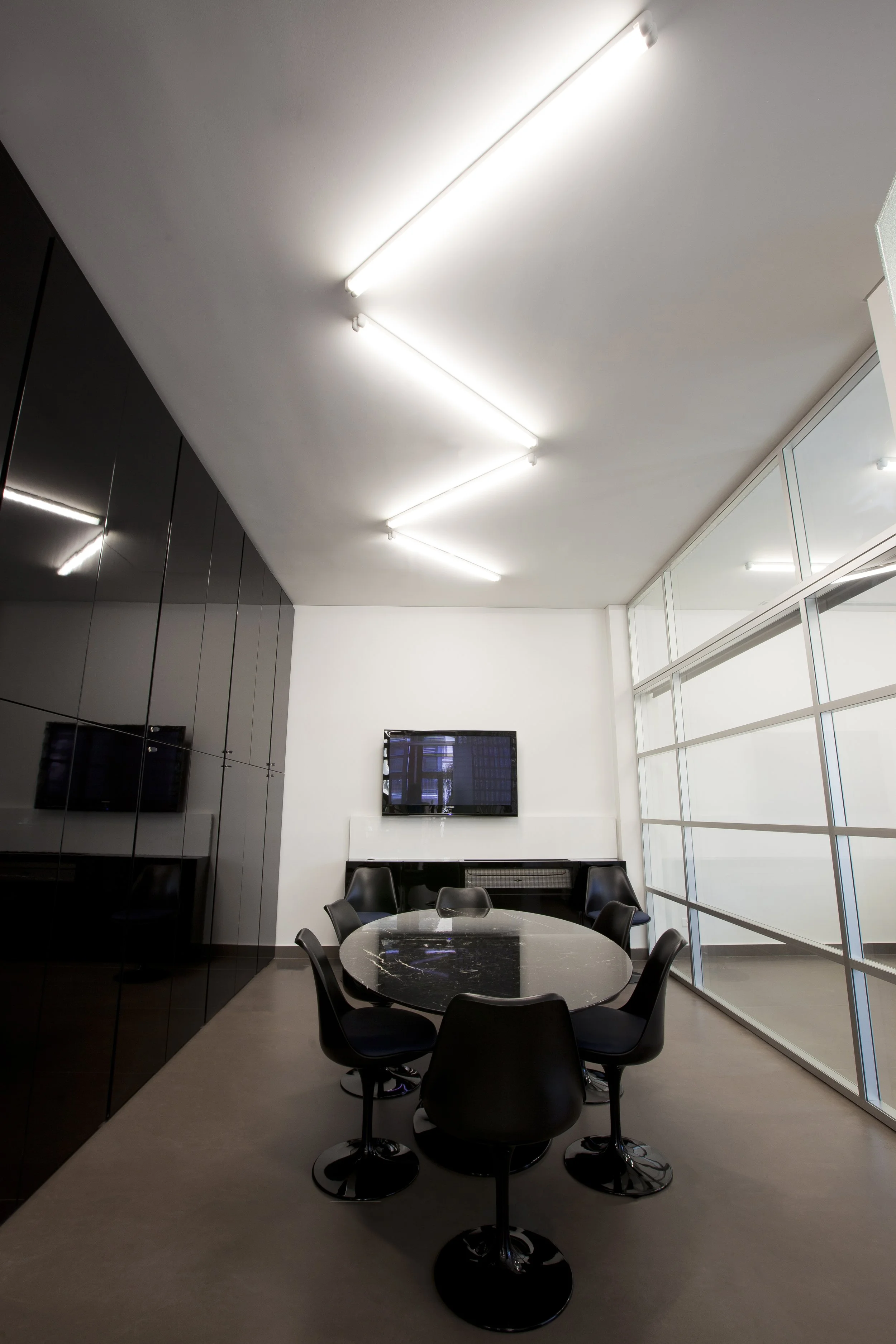 Modern conference room with a round black marble table surrounded by six black chairs, a large flat-screen TV mounted on the white wall, and a glass wall on the right side letting in natural light. The ceiling has white fluorescent lighting.