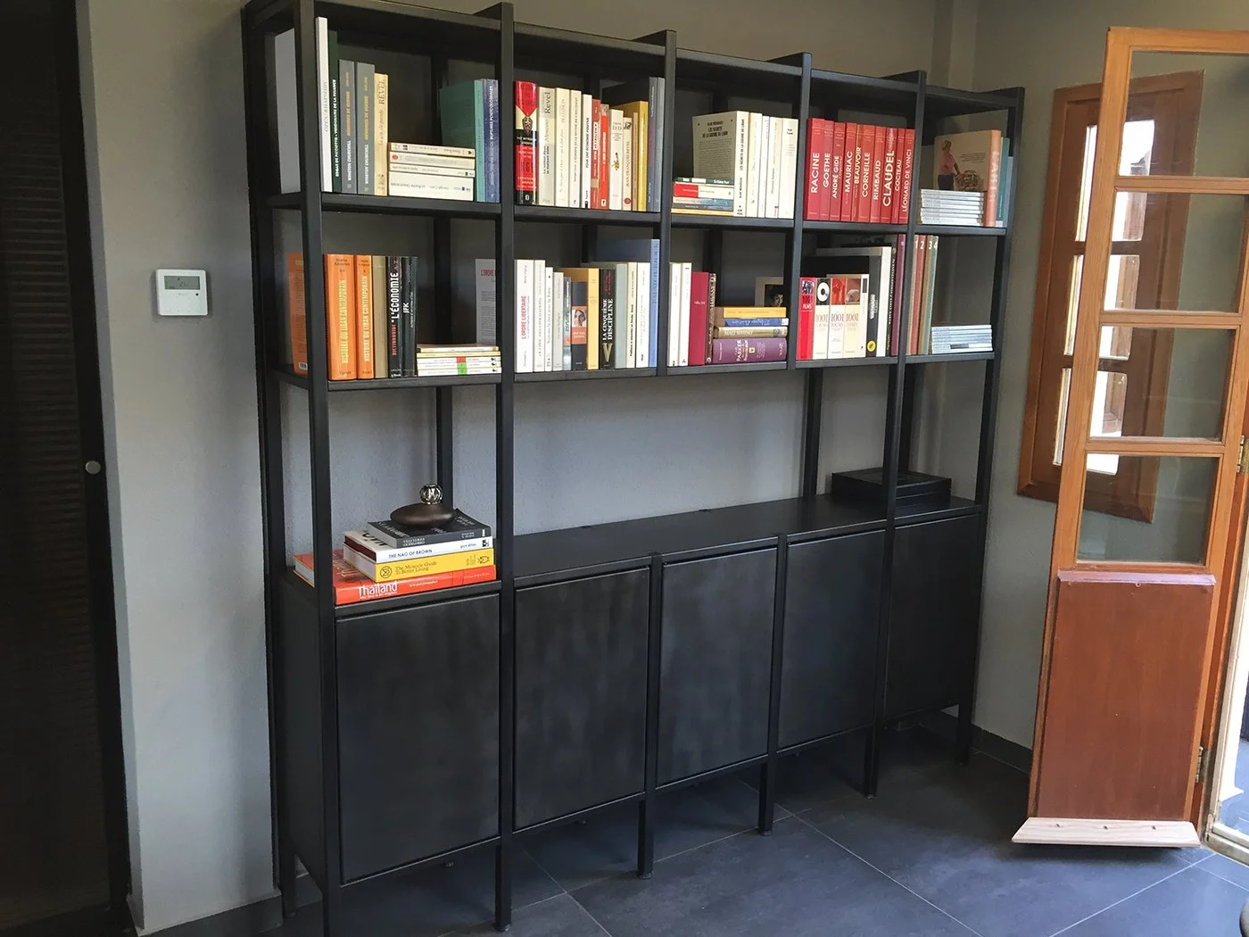 A black metal bookshelf filled with books in a room near a wooden door with glass panels, a gray wall, and a wall-mounted thermostat.