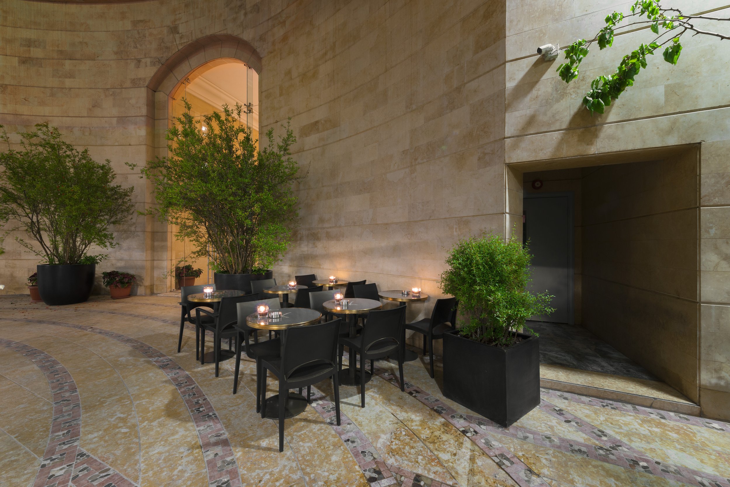Outdoor patio area with black tables and chairs, potted green plants, and candles, surrounded by a beige stone wall with arched window and green vine.