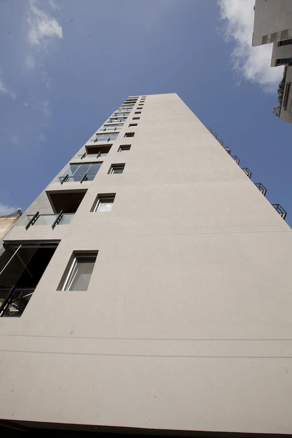 Low-angle view of a modern high-rise apartment building with glass balconies, set against a blue sky with clouds.