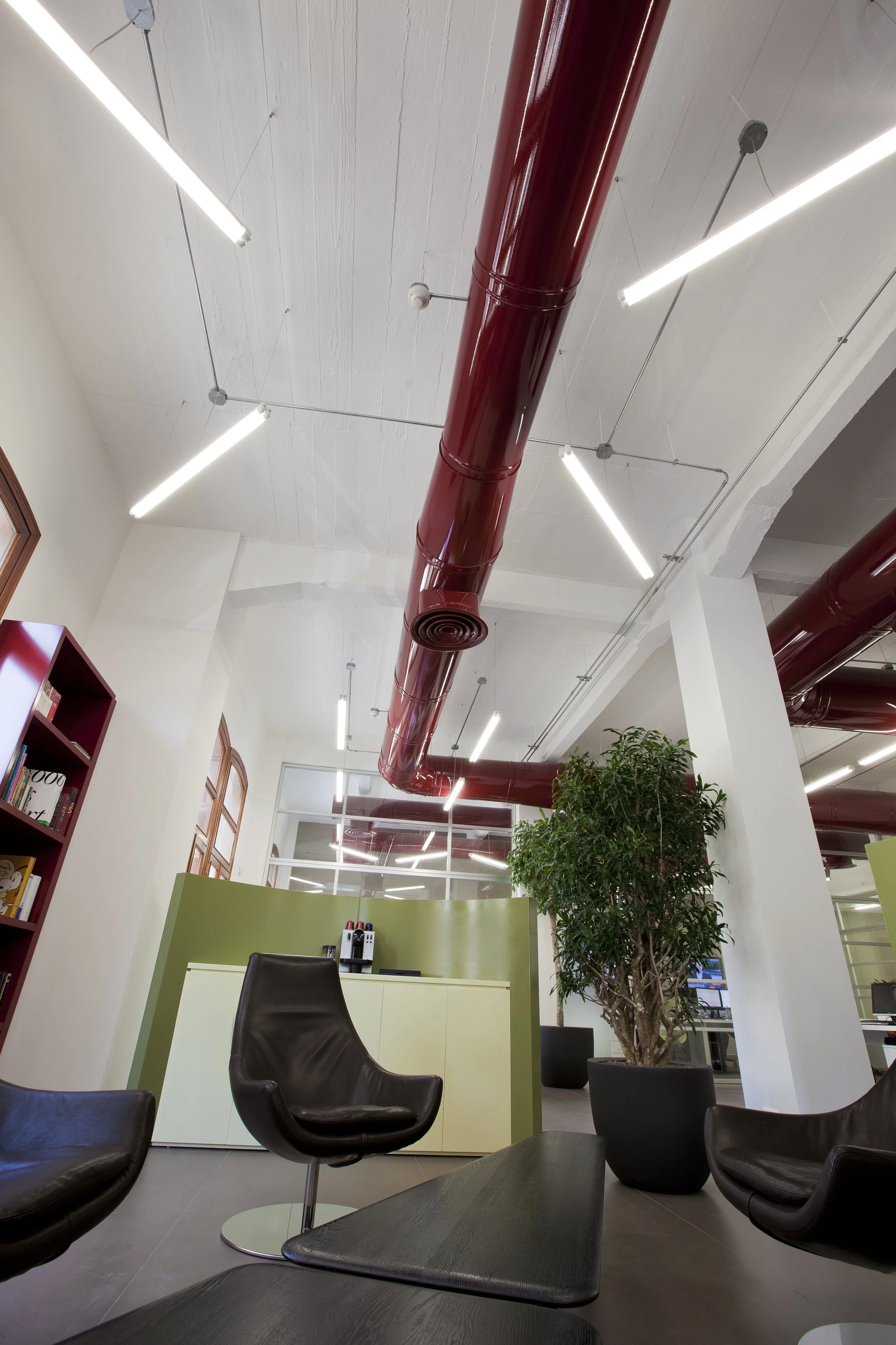 Modern office lounge area with black leather chair, green partition, large potted plant, and red overhead piping with ceiling lights.