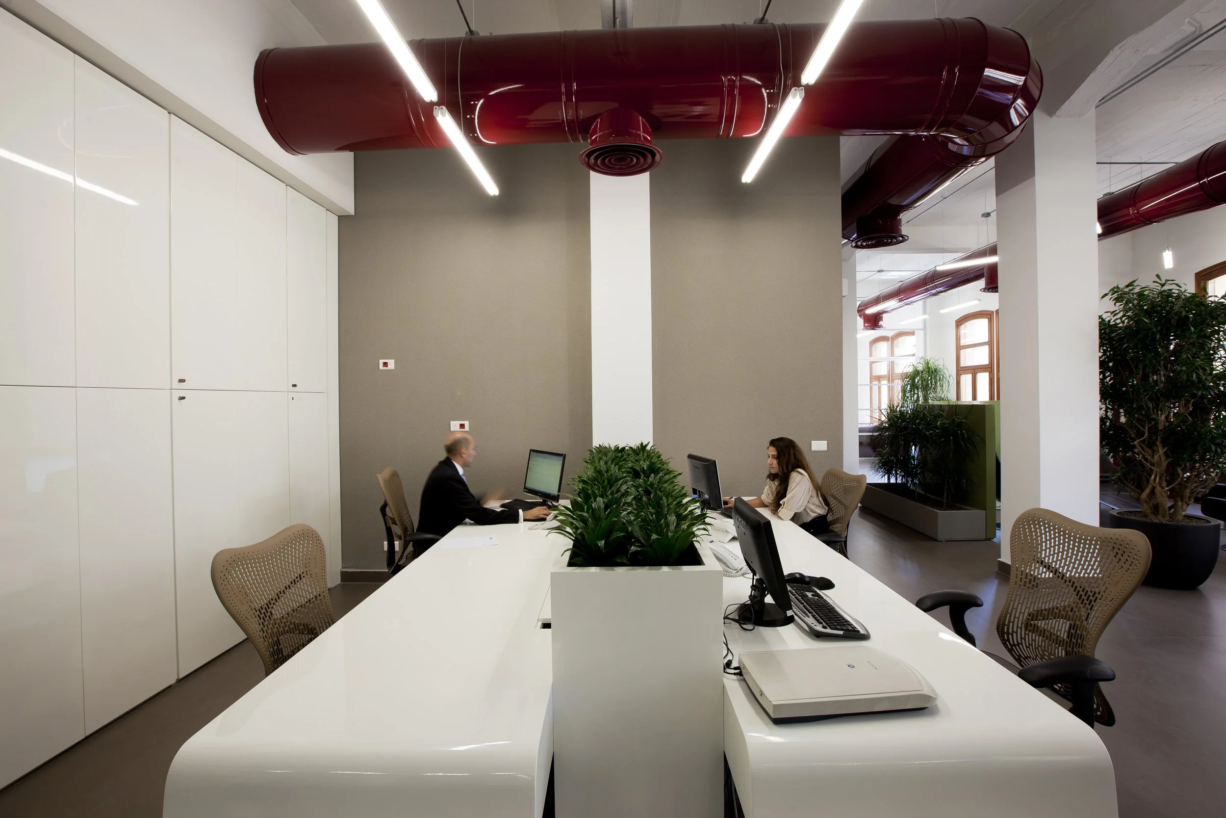 Modern office space with two employees sitting at a white conference table, working on computers. The room features large windows with wooden frames, several indoor plants, and a ceiling with red ducts and built-in lighting.