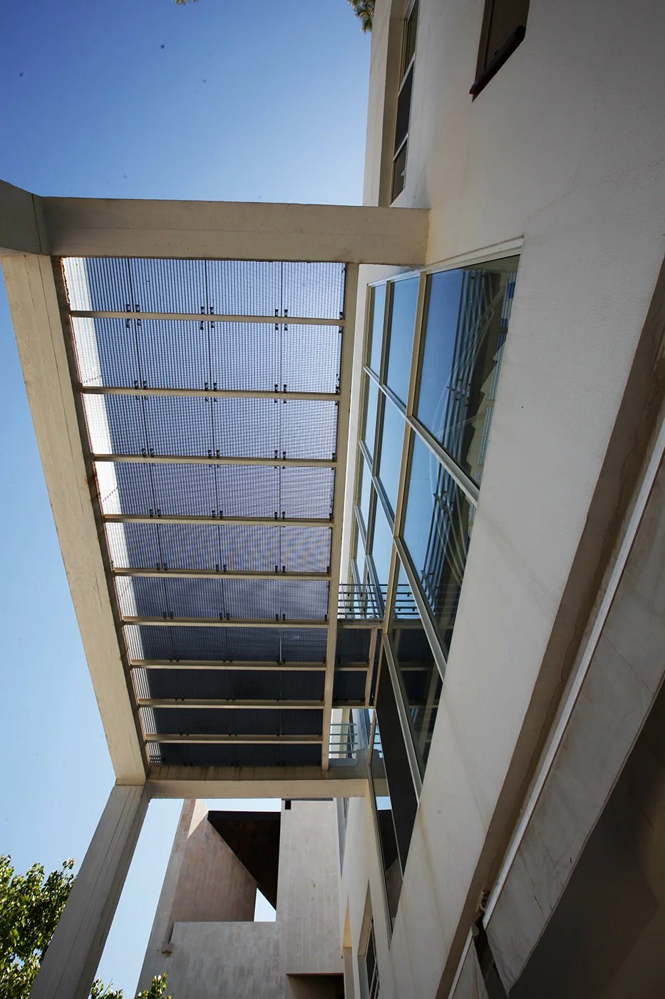 View of modern building exterior with glass windows, white concrete walls, and an overhanging structure with a metal grid roof, under a clear blue sky.