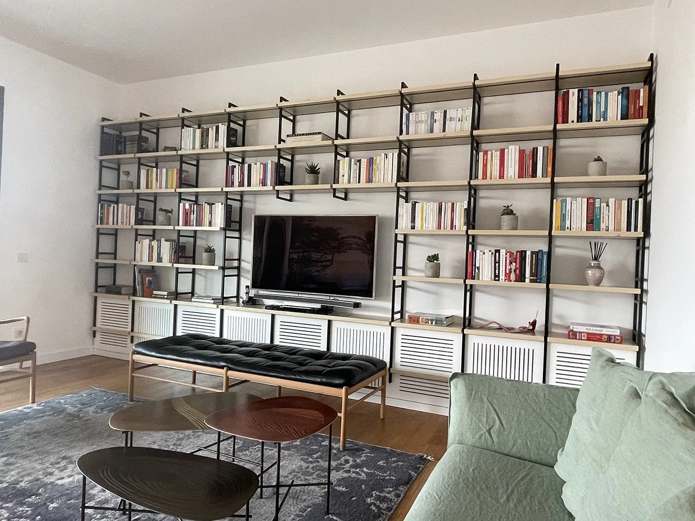 Living room with a large black and white bookshelf wall, a flat-screen TV, a green armchair, a black bench, and nested coffee tables on a patterned rug.