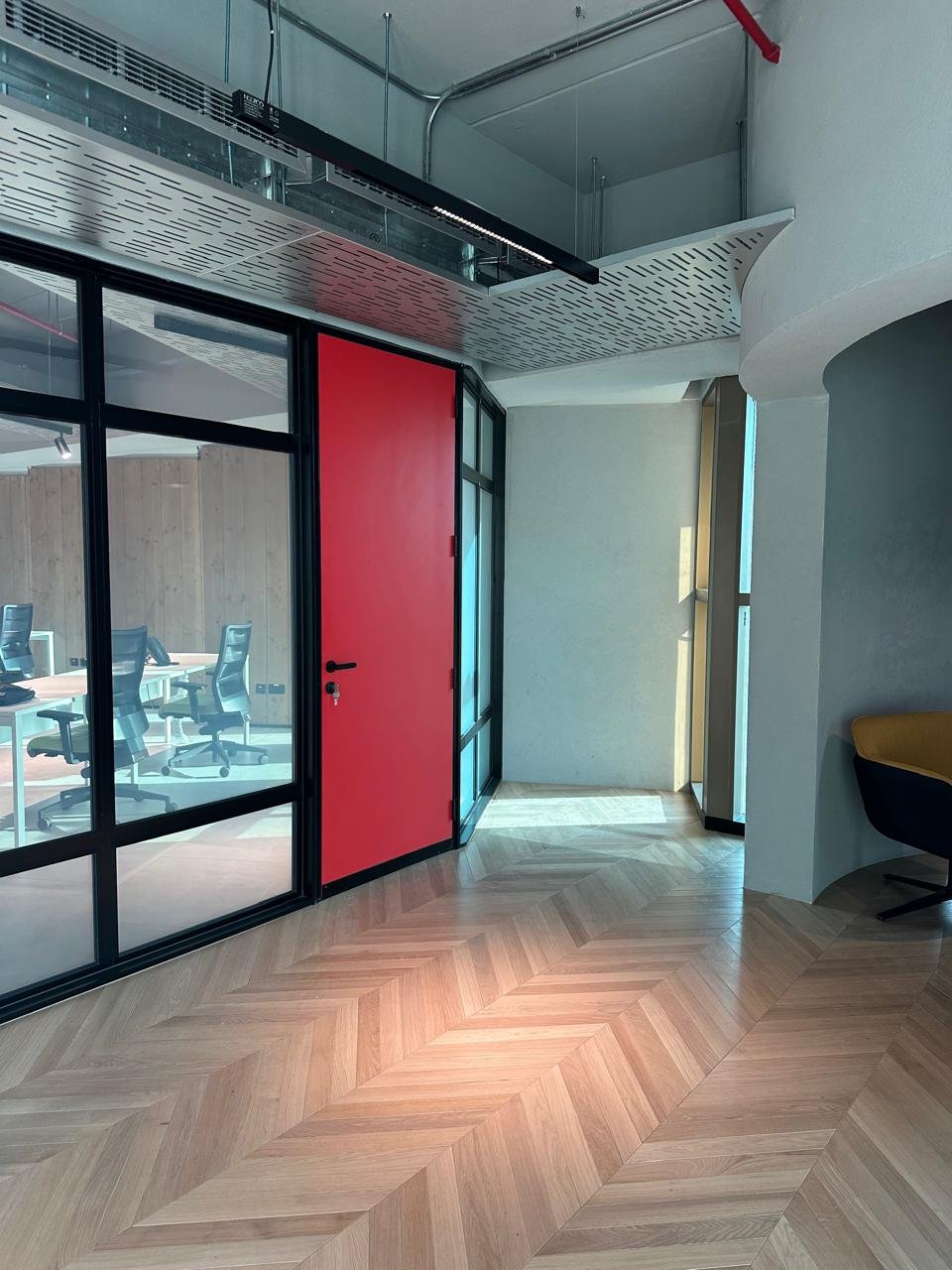 Modern office interior with a glass wall, a red door, wooden herringbone flooring, and a sitting area with a yellow and black chair.