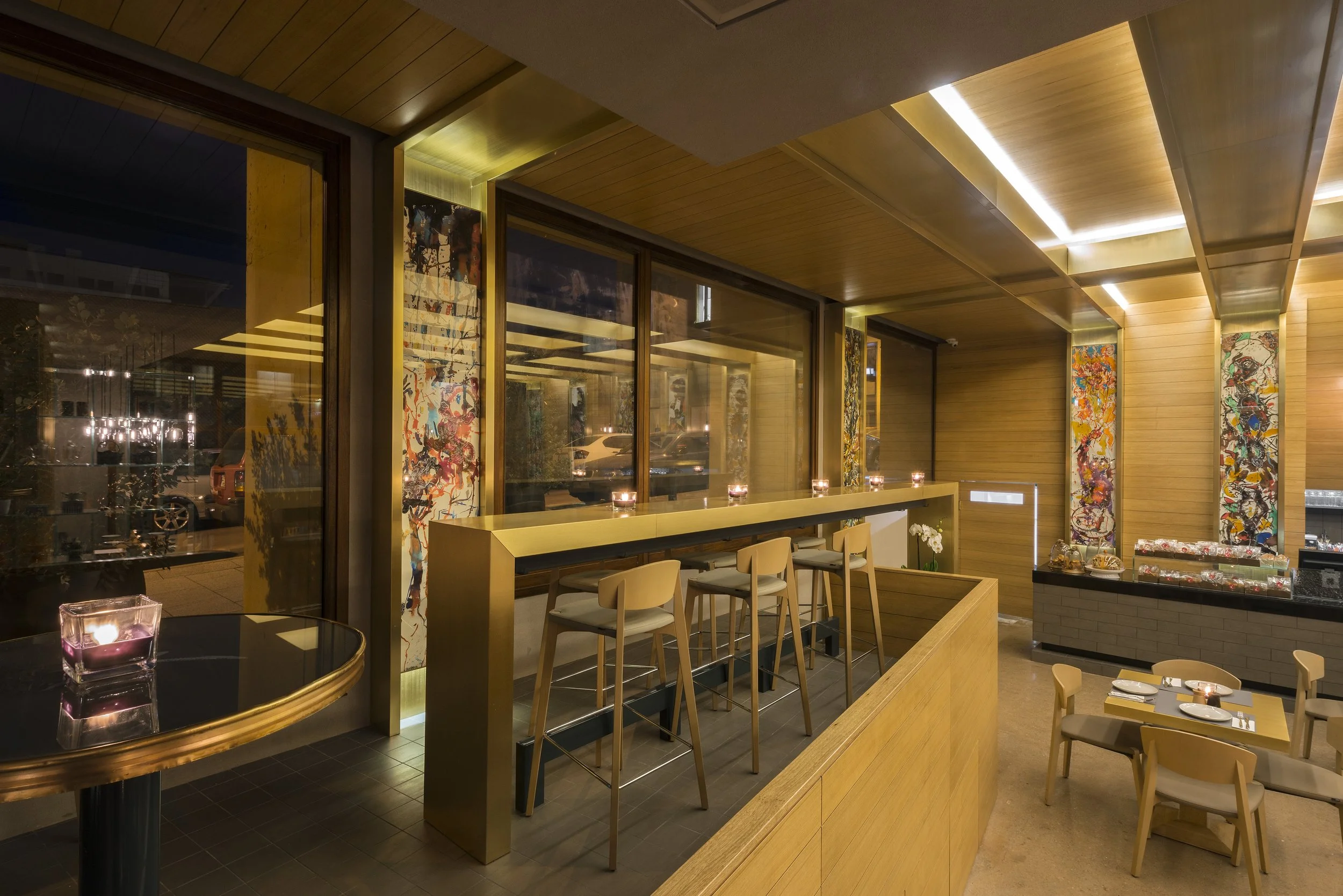 Interior of a modern restaurant with light wood walls, artwork, and ambient lighting. There are chairs at a counter and tables with place settings, candles, and decorative items.