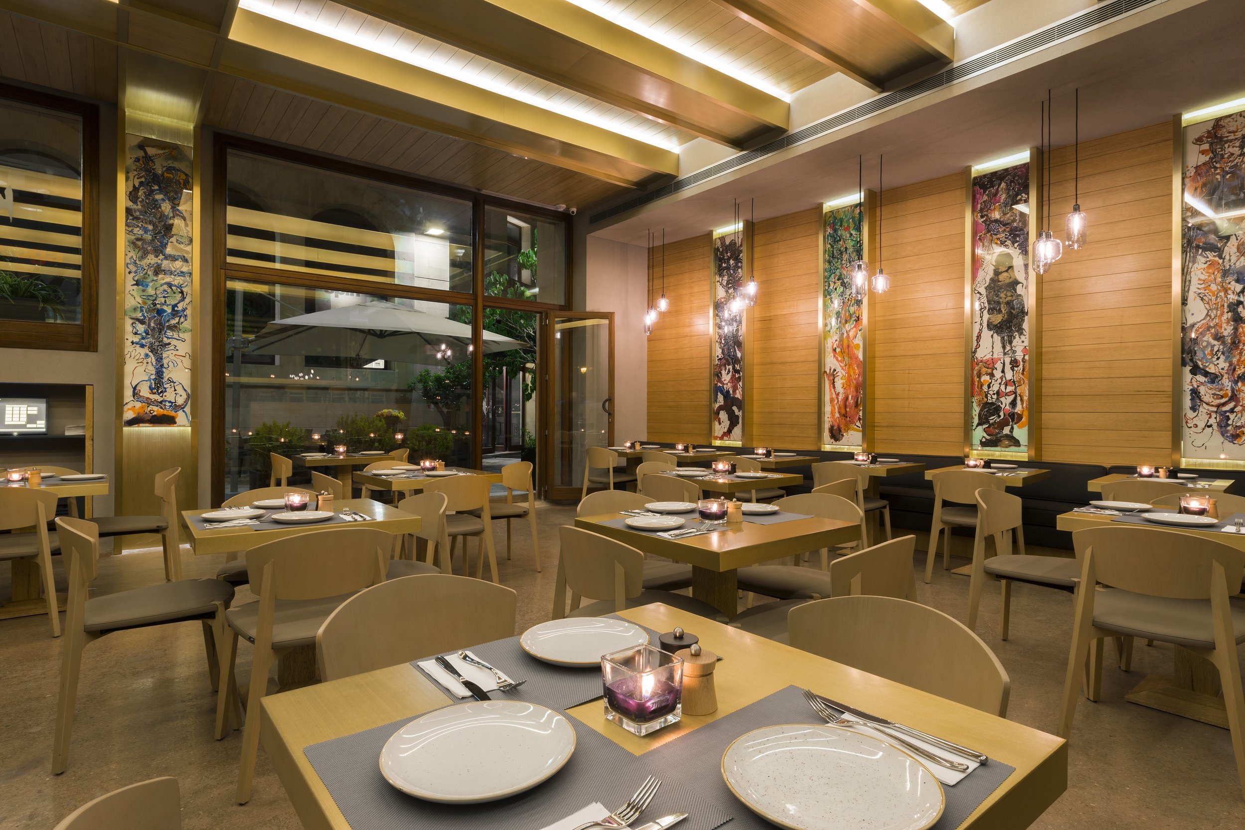 A modern restaurant interior with wooden walls, abstract artwork hanging vertically, and pendant lighting. Tables are set with plates, cutlery, napkins, and purple candles, with large windows showing outdoor greenery.