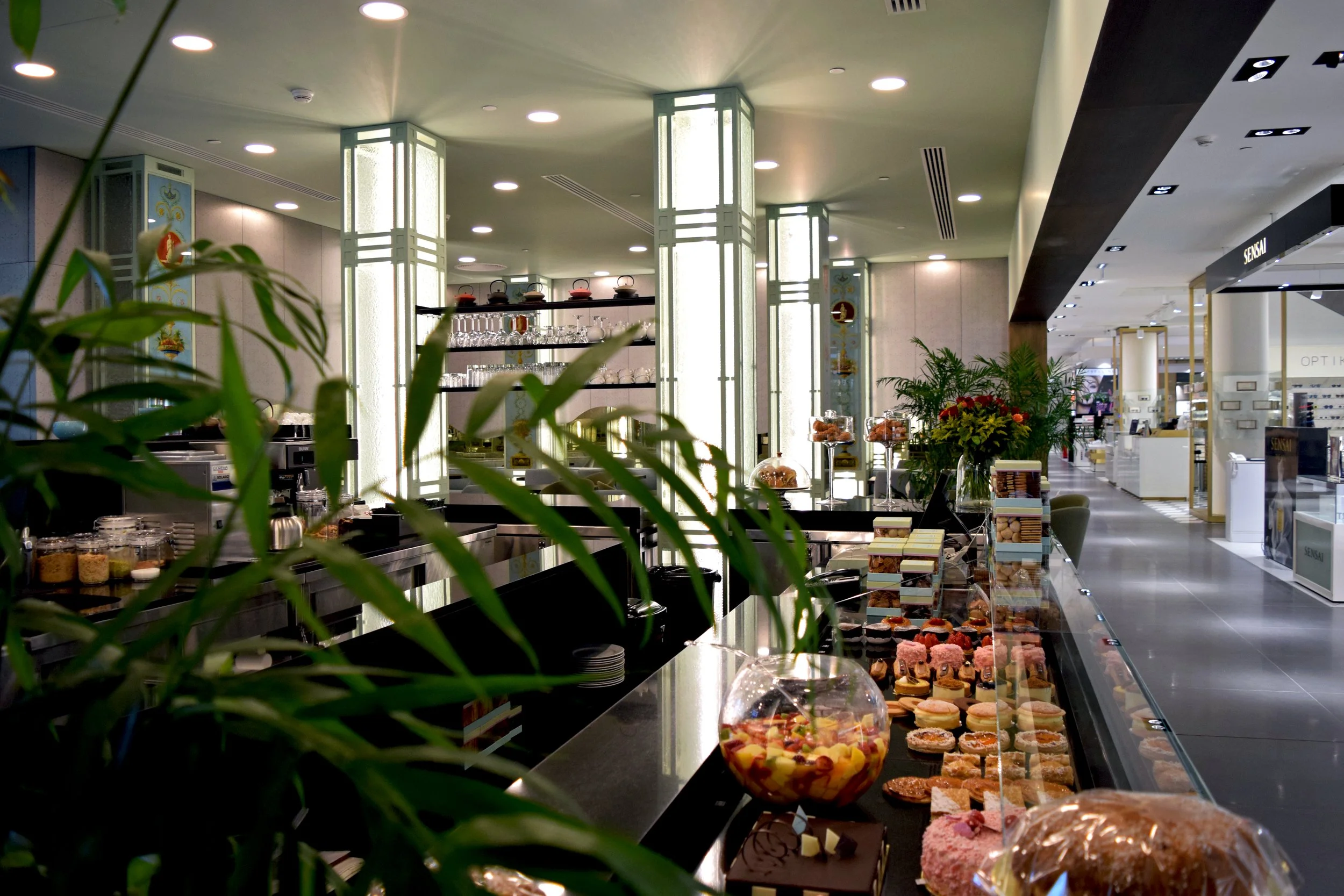 Interior of a bakery or café with display cases filled with pastries, desserts, and baked goods. There are decorative plants and illuminated pillars, with drinking glasses and teapots on shelves in the background.