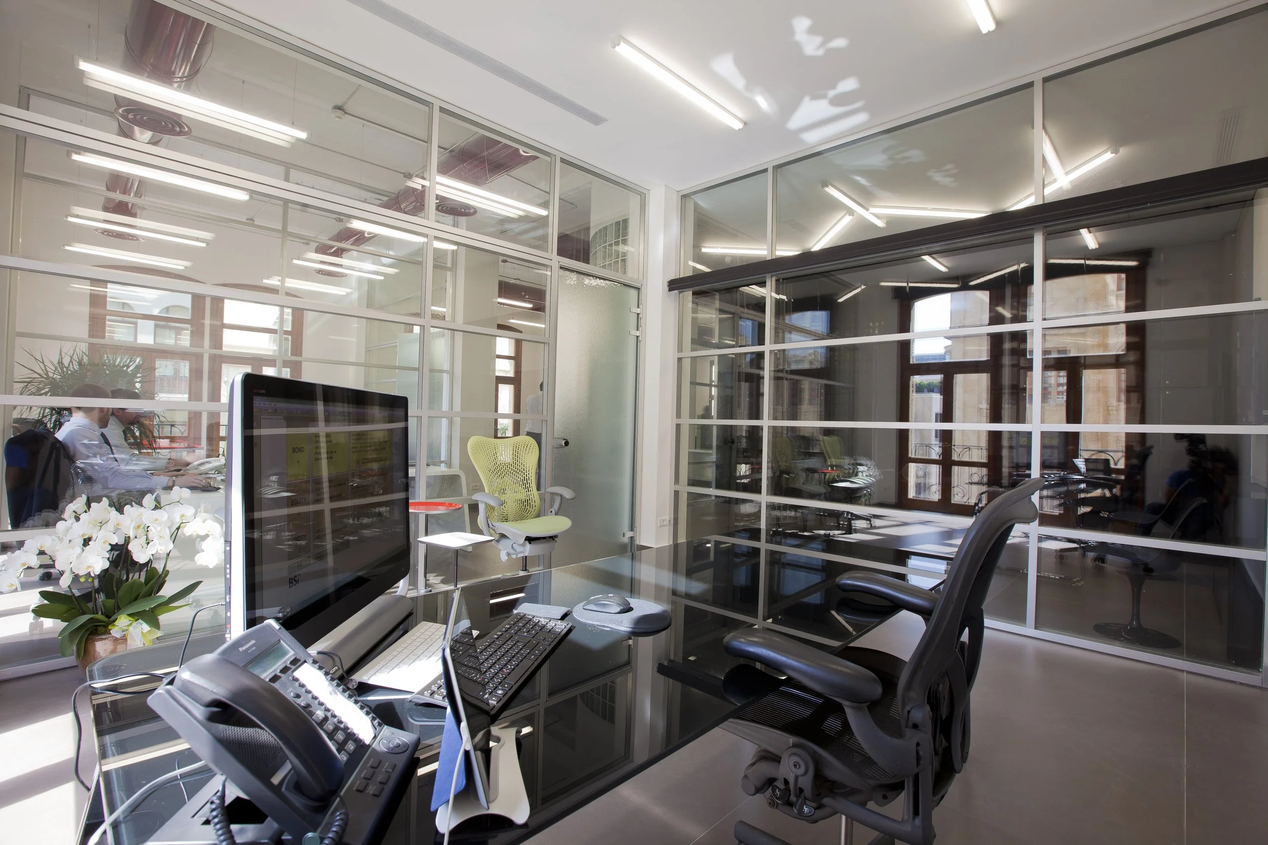 Modern office space with glass partitions, desks, computers, and office chairs, illuminated by fluorescent ceiling lights. There is a desktop monitor, a keyboard, and a telephone on one desk, with a plant and white orchids nearby. Two people are working in the background.