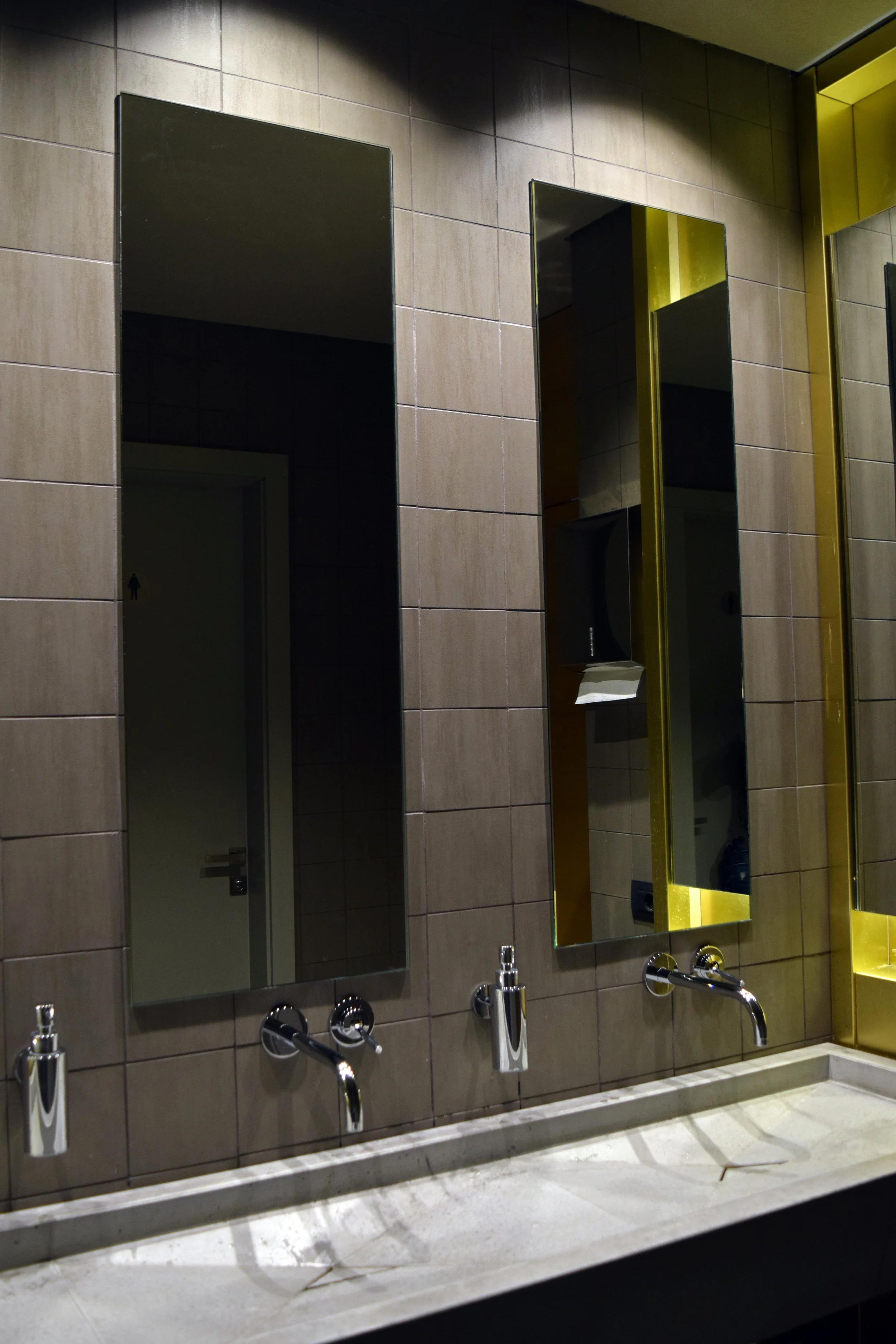Public restroom with two mirrors and two sinks, tiled walls, soap dispenser, and yellow accents on the wall.