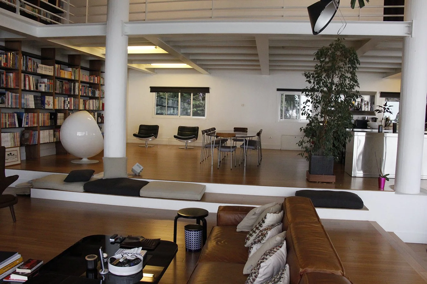 A modern, open-concept living space with a brown leather sofa, a black coffee table, a white built-in bench with cushions, a large potted plant, and a dining area with a round table and six chairs. There's a bookshelf filled with books, large windows
