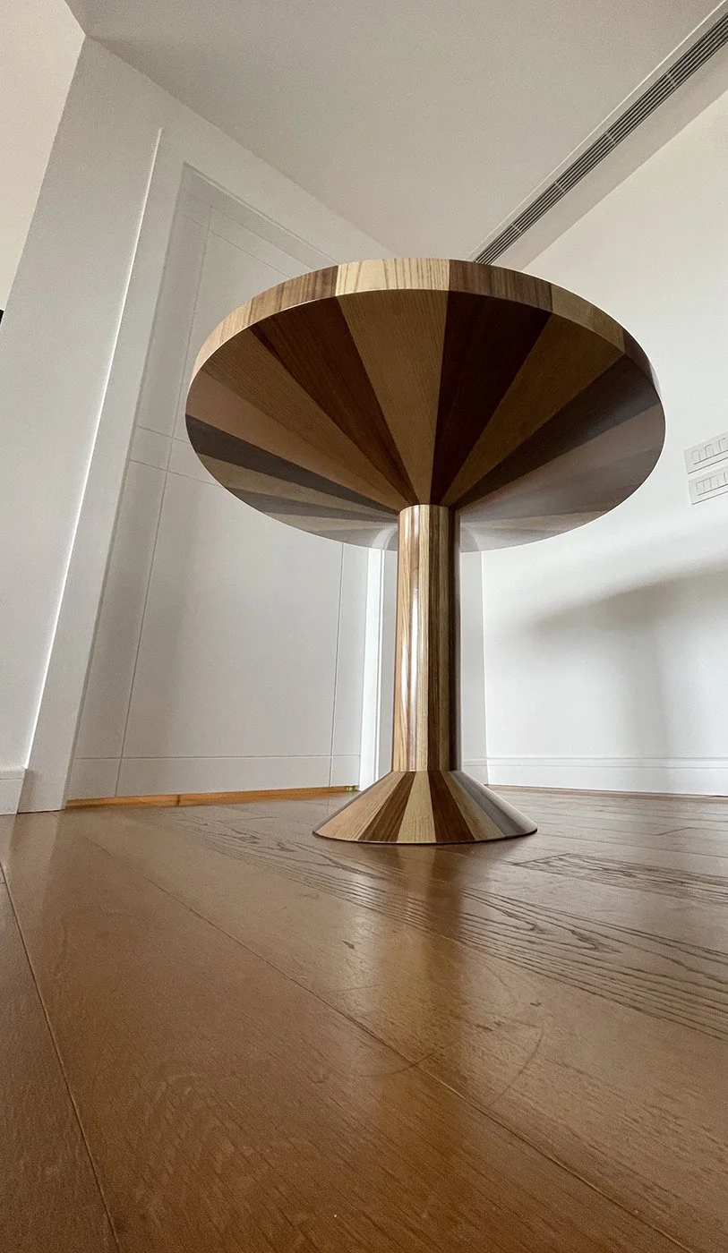 A modern wooden side table with a round top and a single central support, featuring a geometric wood pattern in various shades. The table is on a wooden floor in a minimalistic room with white walls.