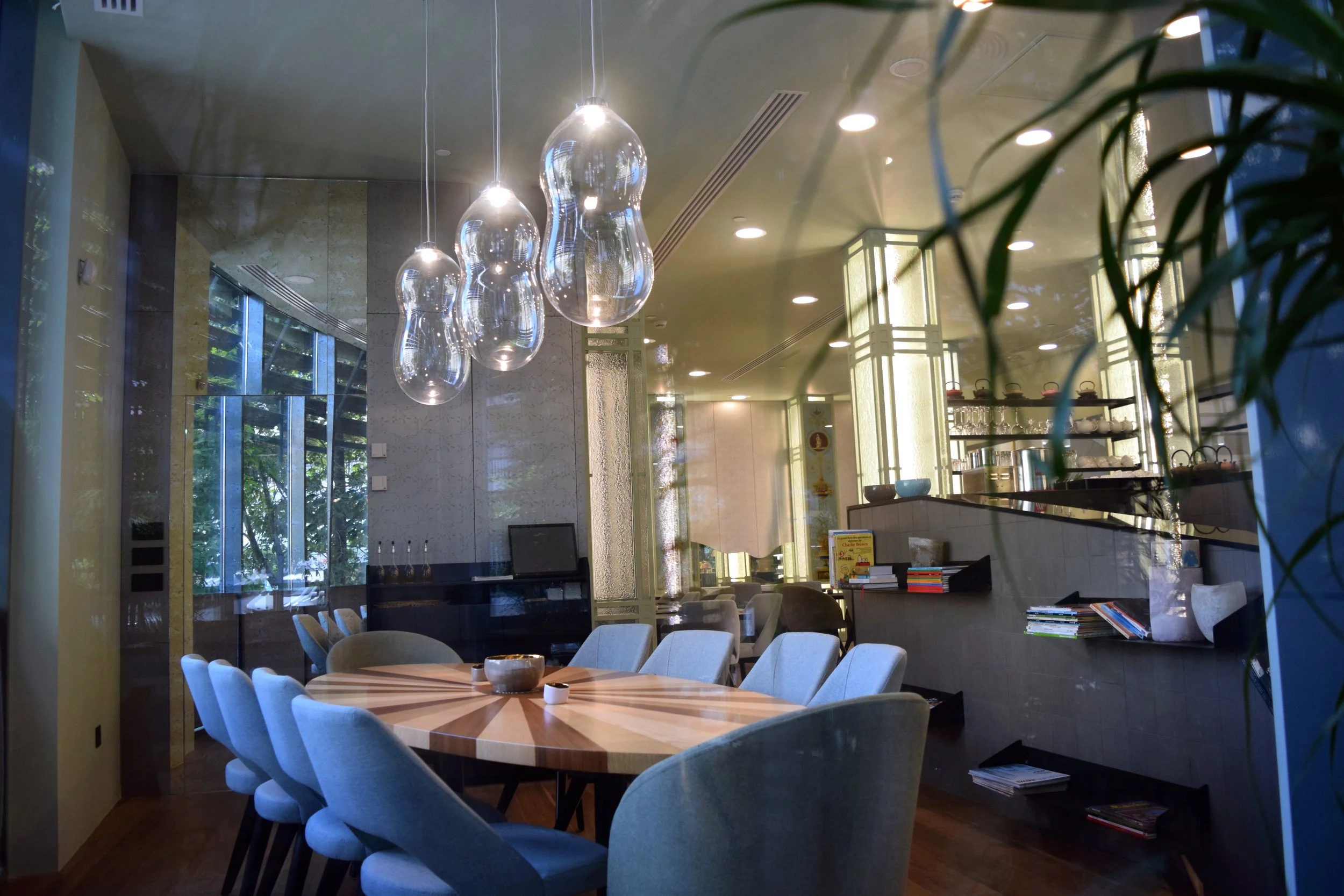 Modern restaurant interior with a large wooden table surrounded by light blue upholstered chairs, sleek glass pendant lights hanging from the ceiling, books and decorative items on a black table, large windows with blinds, and lush green foliage outs