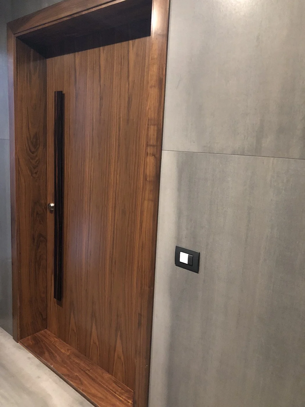 Wooden door with vertical black handle, surrounded by gray wall panels and a black light switch.