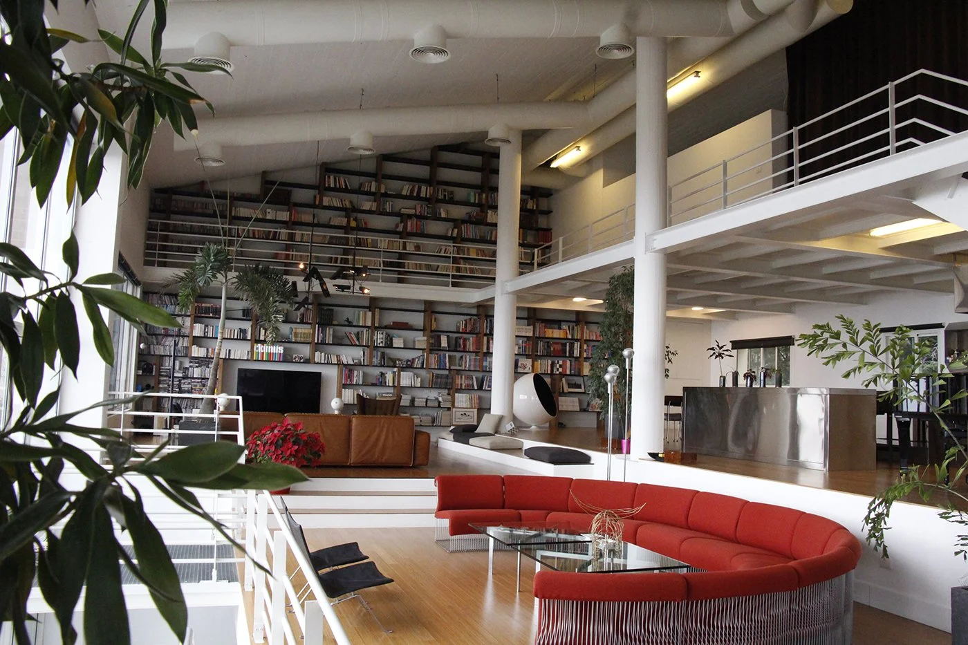 Spacious modern living room with curved red sofa, glass coffee table, large bookshelf wall, and indoor plants.