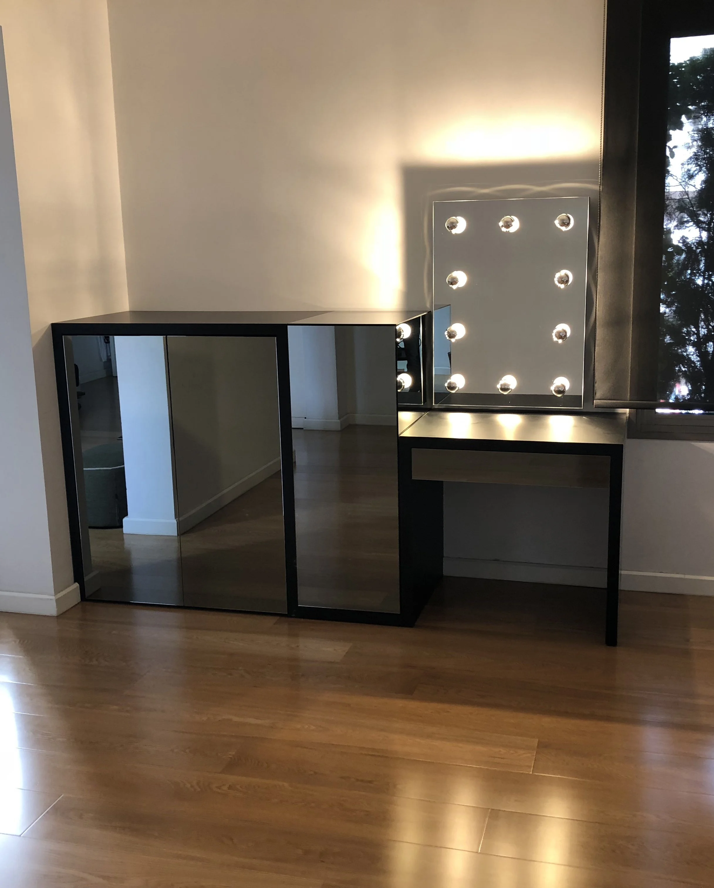 Interior space with hardwood flooring, a mirrored vanity table with lights, and a window with a view of trees outside.