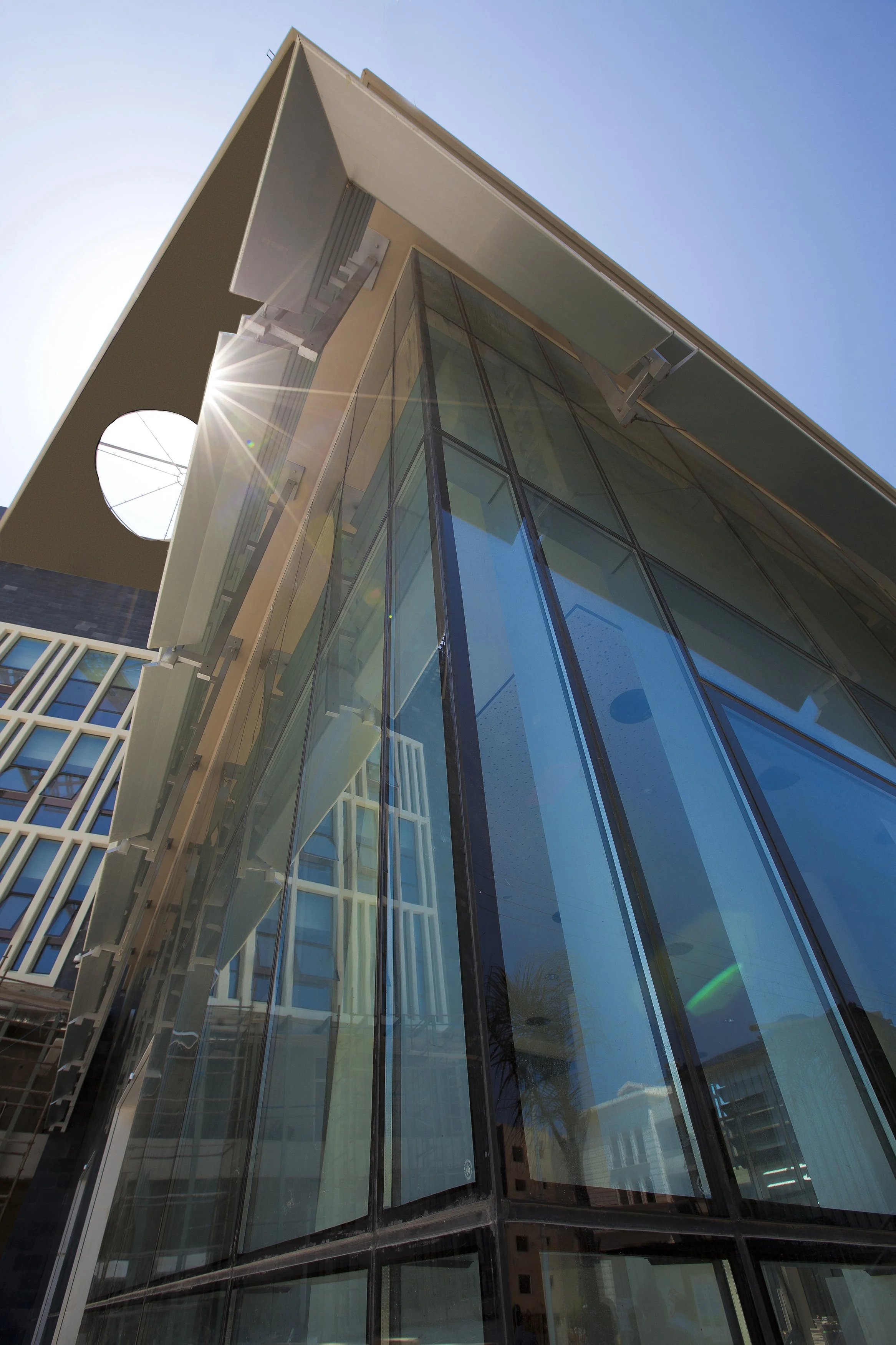 Side view of a modern multi-story building with glass windows and concrete structure under a clear blue sky. The sunlight creates a lens flare effect.