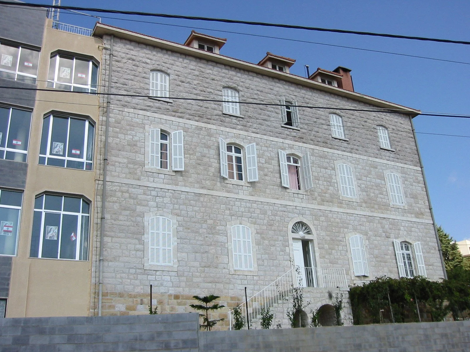 Multi-story building with brick facade, white arched window shutters, and a door with a rounded fanlight, set on a small landscaped hill with plants and steps.