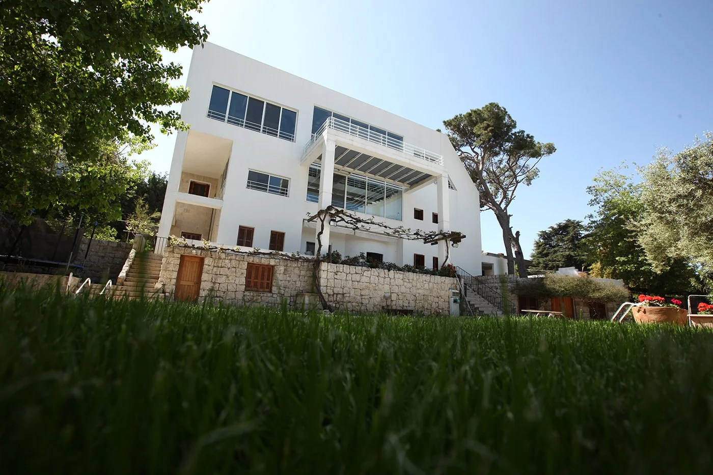 Modern white multi-story house with large windows, surrounded by trees and greenery, with a stone wall and outdoor stairs.
