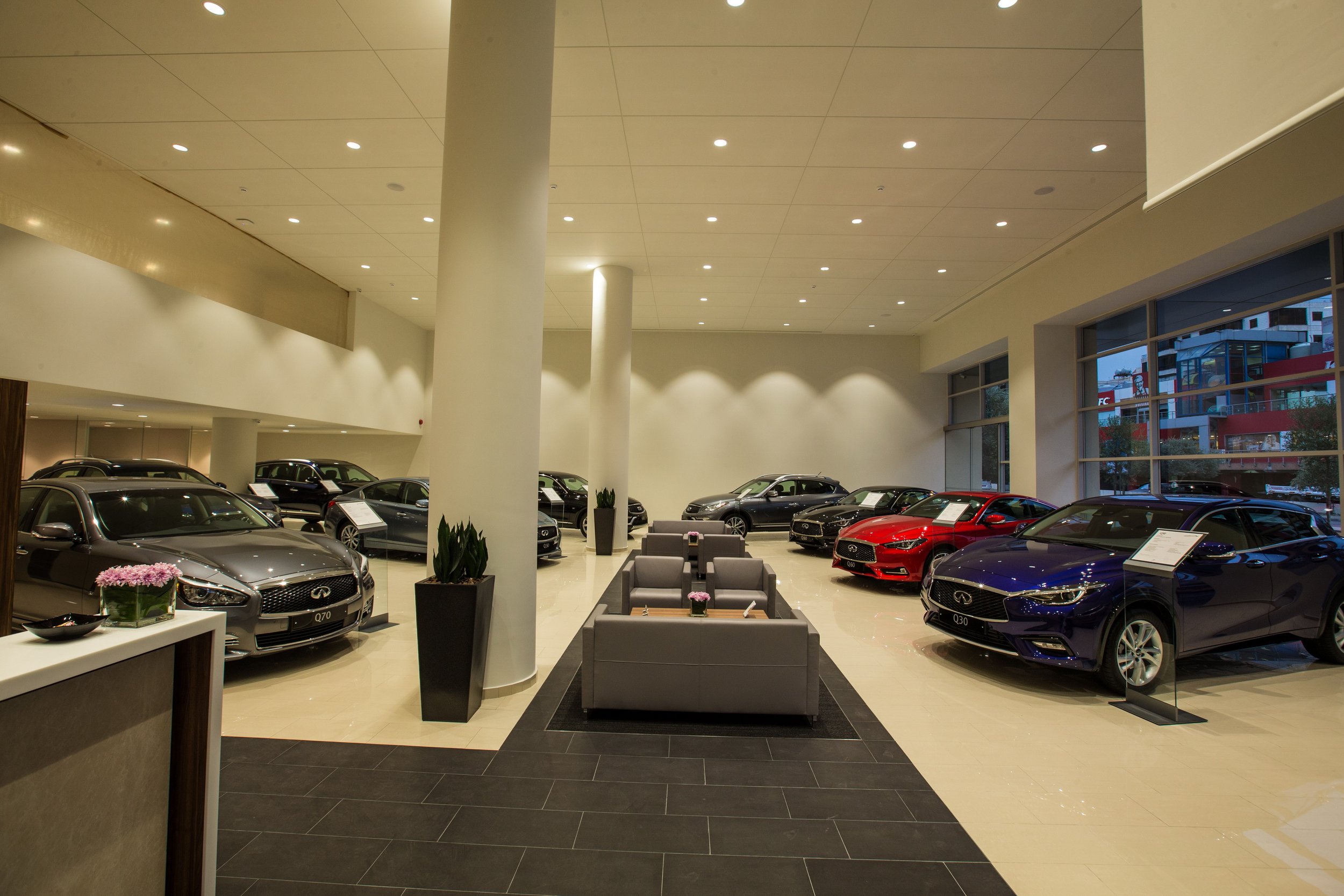 Interior of a luxury car dealership showroom with multiple parked cars, modern seating area, large windows showing buildings outside, and bright ceiling lights.