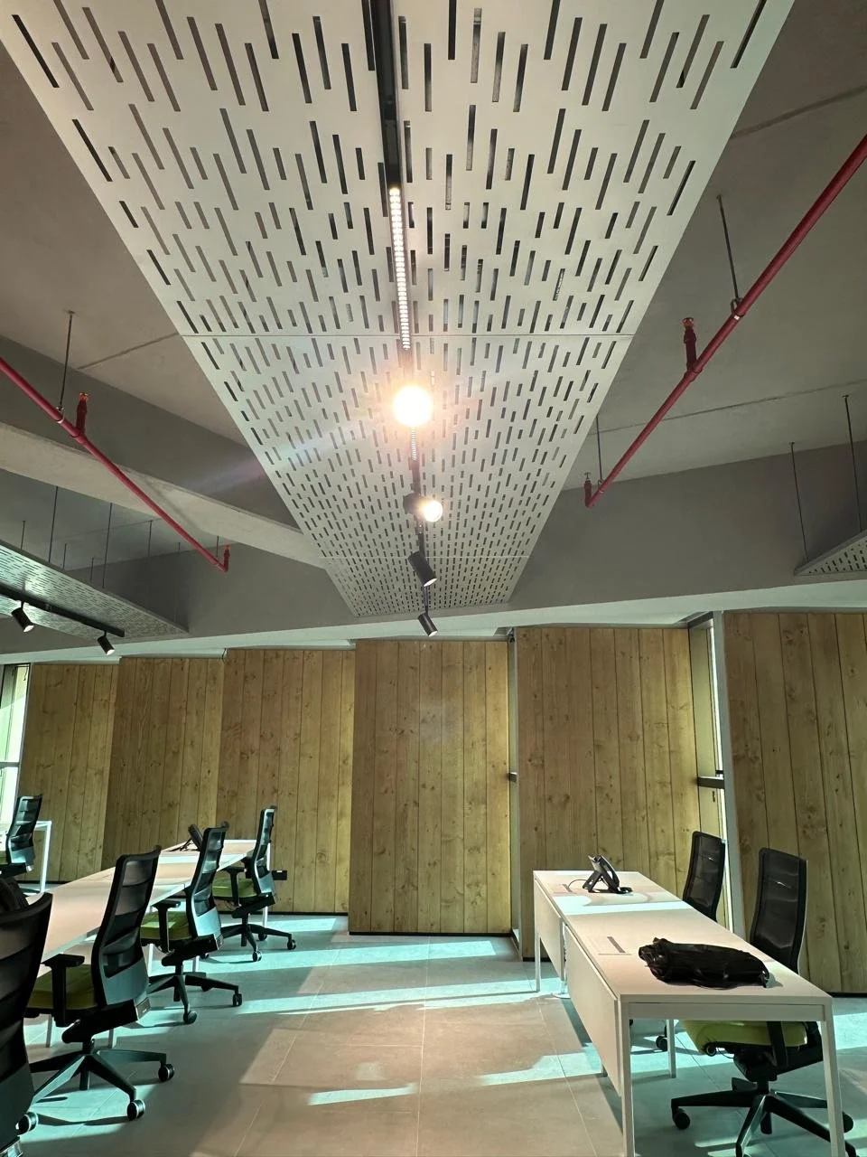 An office conference room with a wooden wall, white desks, black office chairs, a phone, and a black bag, lit by ceiling lights