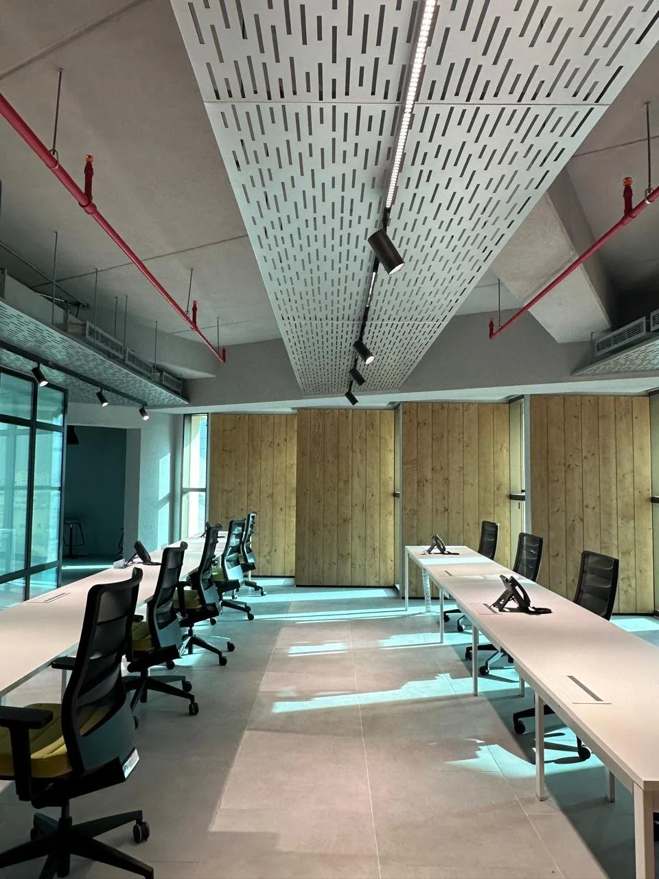 Empty modern conference room with white tables, black chairs, wooden wall panels, and large windows, illuminated by natural light and ceiling track lighting.