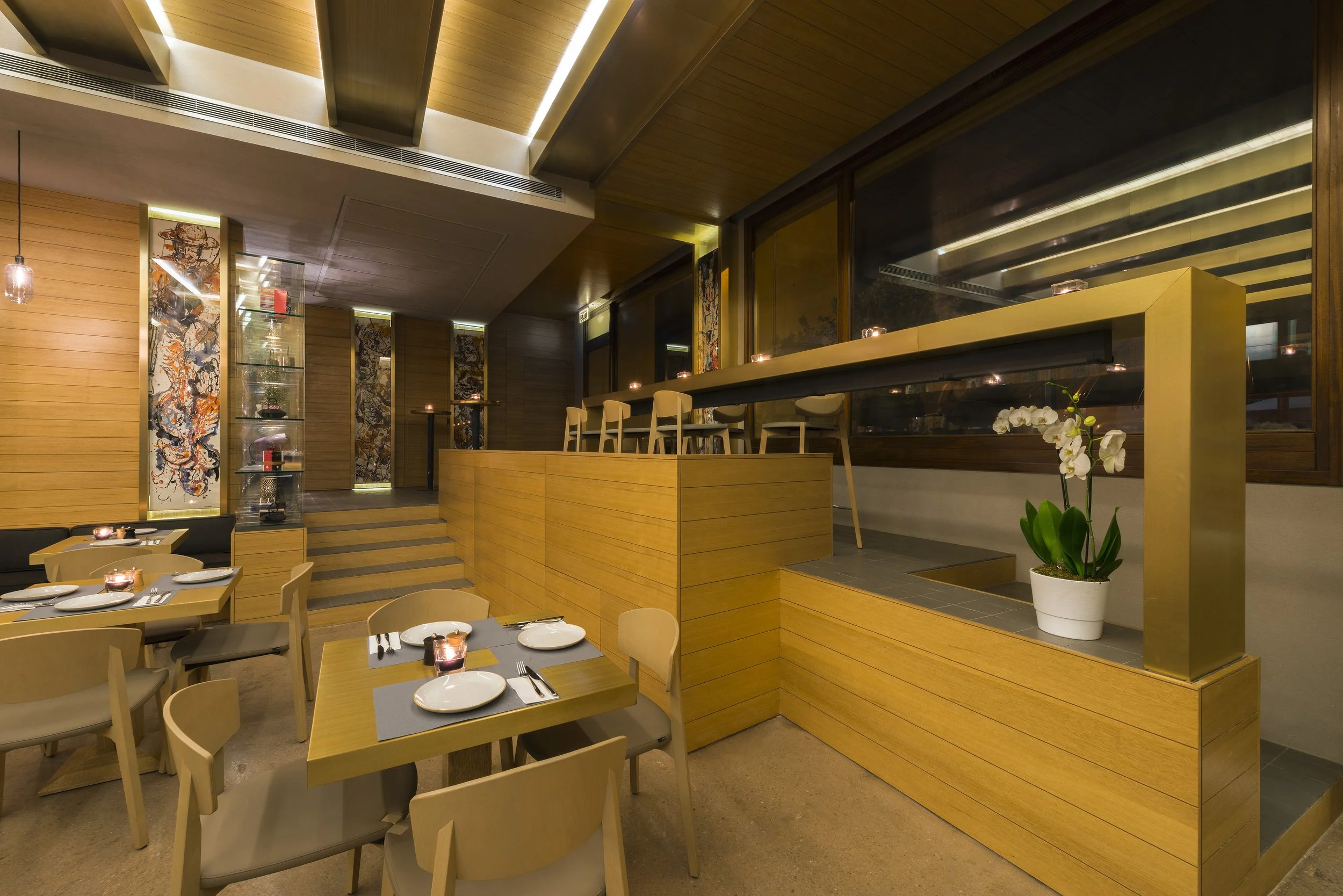 A modern restaurant interior with wooden walls and tables set with plates, forks, knives, and candles, illuminated by warm lighting, with a large window and decorative art panels.