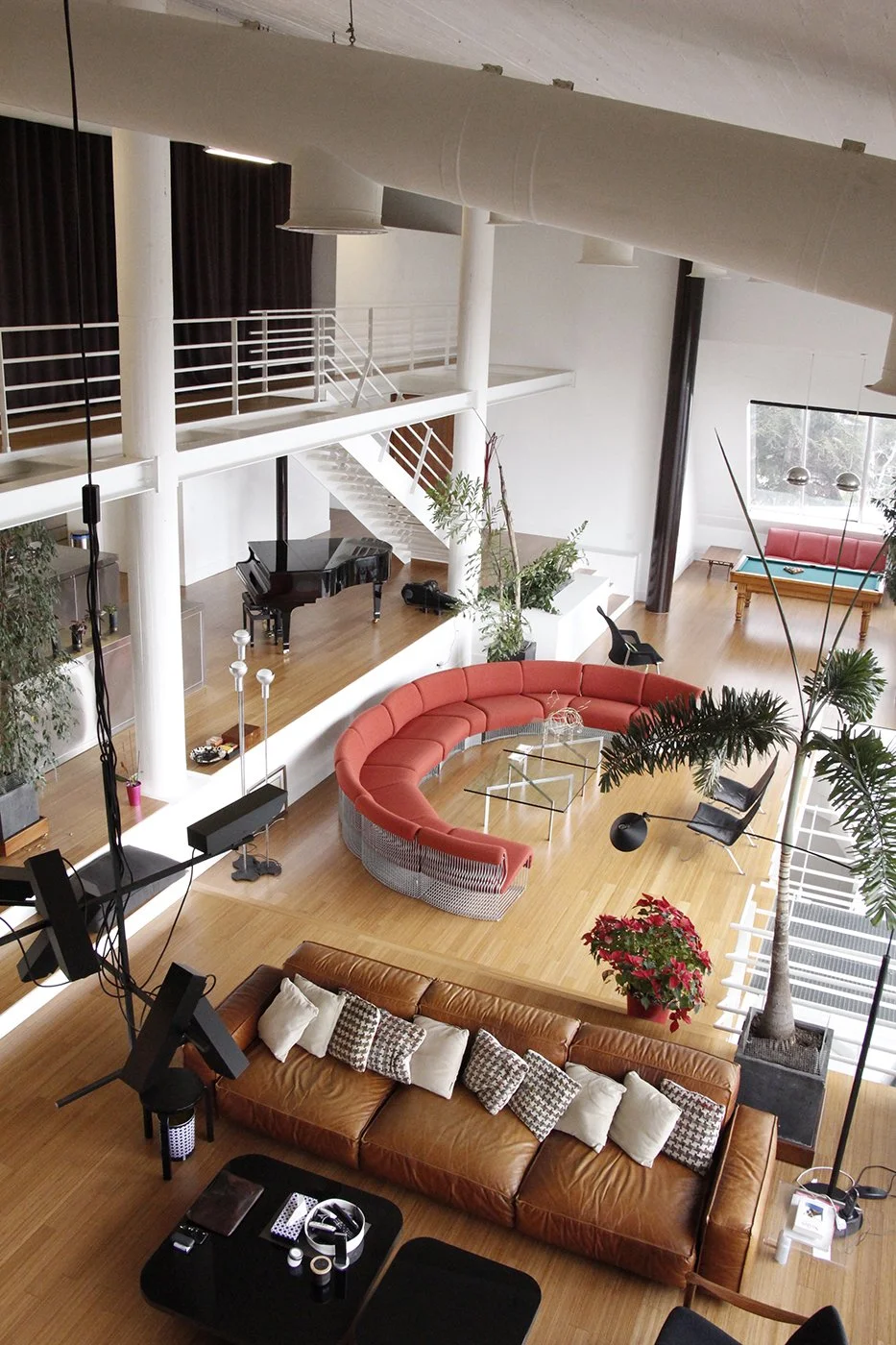 A spacious, modern living room with wooden flooring, white walls, and large windows. It features a curved red sofa, a brown leather couch with pillows, a black grand piano, and various chairs. There are tall indoor plants, a staircase with white rail