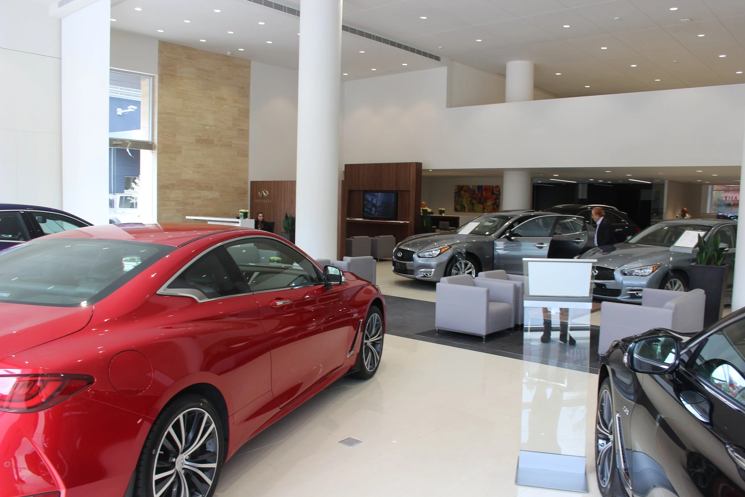 Inside a modern car dealership showroom with several new cars, including a red car in the foreground and a gray car beside it, and a sales representative talking to a customer in the background.