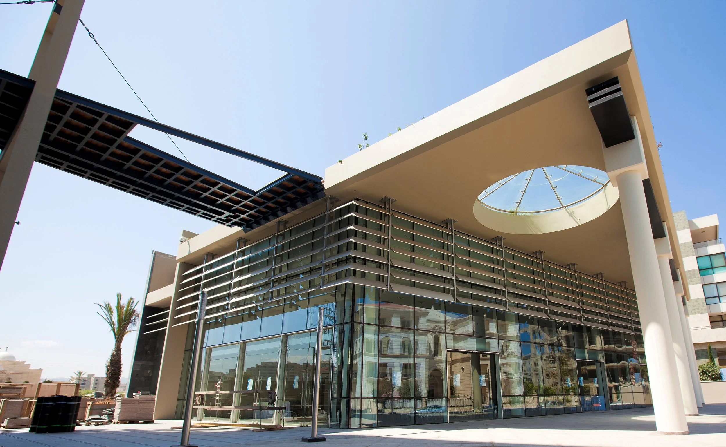 Modern building with glass windows, metal accents, and a circular window on the upper level. Clear blue sky in the background, with a palm tree visible on the left.