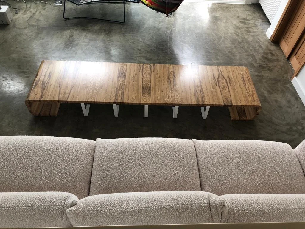 A wooden bench with a natural finish, placed on a concrete floor in front of a beige sofa inside a room.