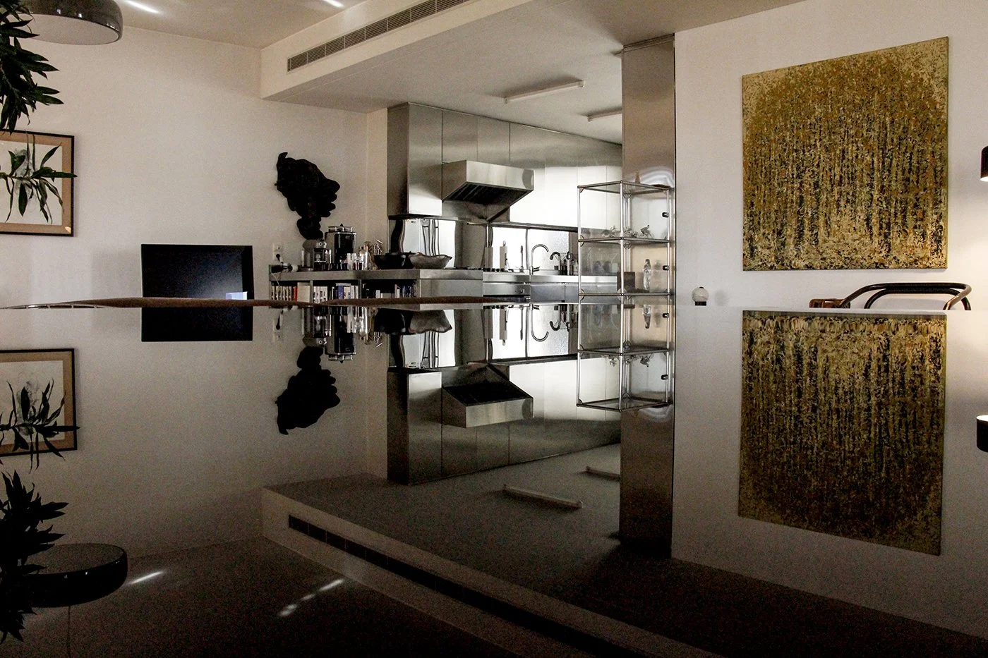 An interior view of a modern kitchen and art gallery, with a reflective stainless steel kitchen area, artworks on the walls, and a black sculpture or object on a shelf. There is a reflection of a wooden ceiling beam on the floor.
