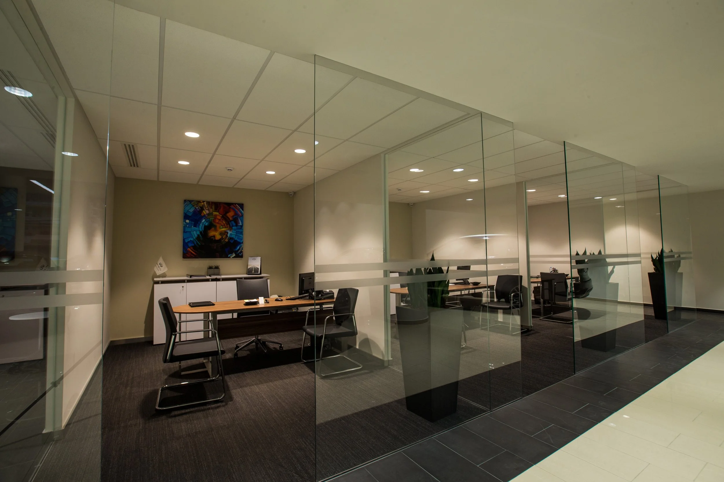 Modern office space with glass partitions, a wooden conference table, black chairs, ceiling lights, a colorful abstract painting on the wall, and potted plants.