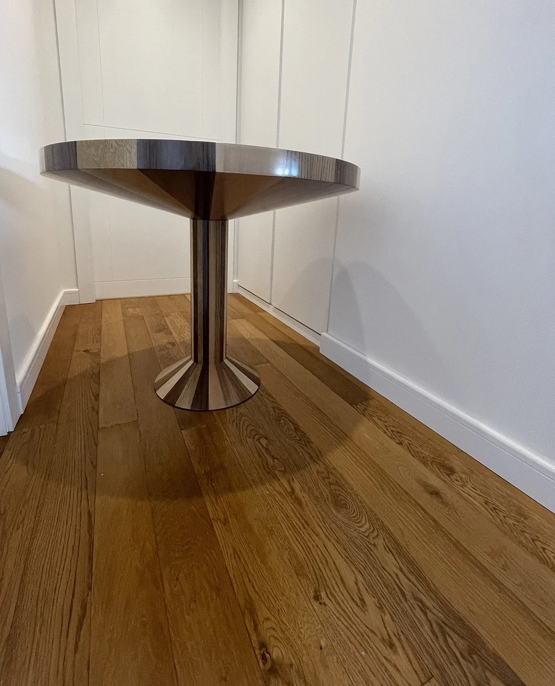 A round metallic table with a pedestal base on a wooden floor in a corner of a room.