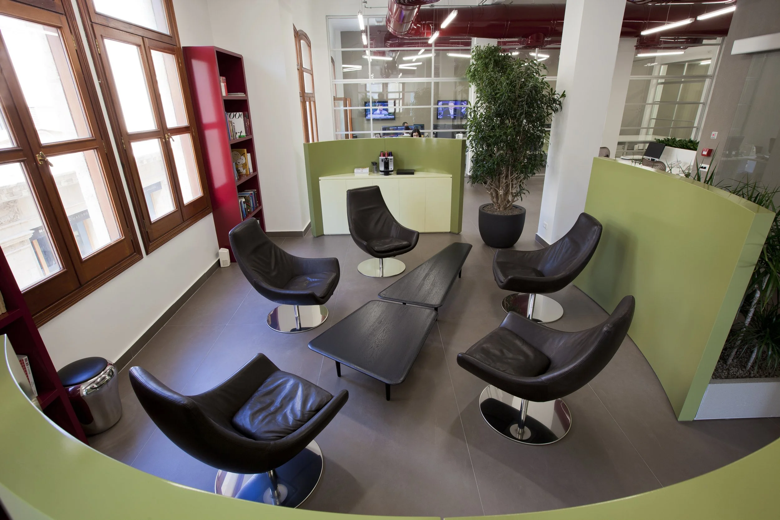 Indoor office lounge area with large windows, black leather chairs, a coffee table, a green partition, a tall potted plant, bookshelves, and a view into a workspace with people working at desks.