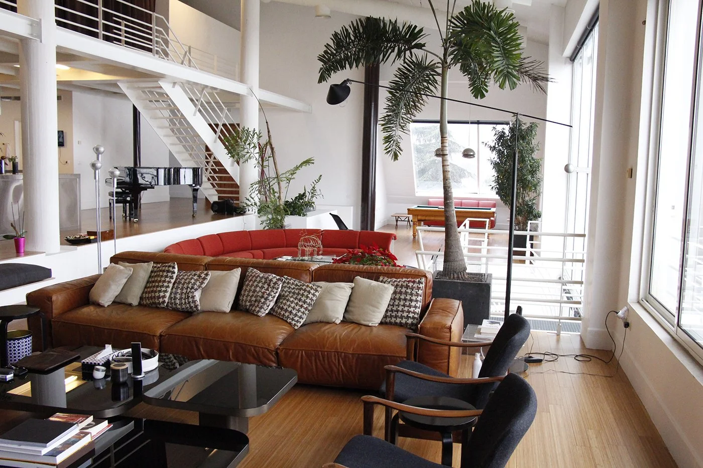 Spacious living room with large windows, leather and fabric sofas, potted plants, and a black grand piano on a raised platform.