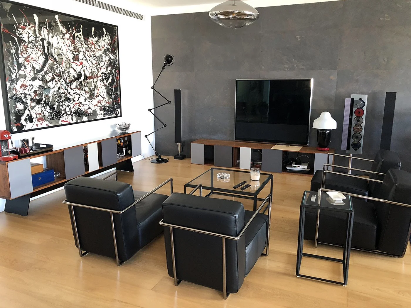 Modern living room with black leather armchairs, a flat-screen TV, abstract artwork, wooden and gray furniture, and decorative items on a light wood floor.