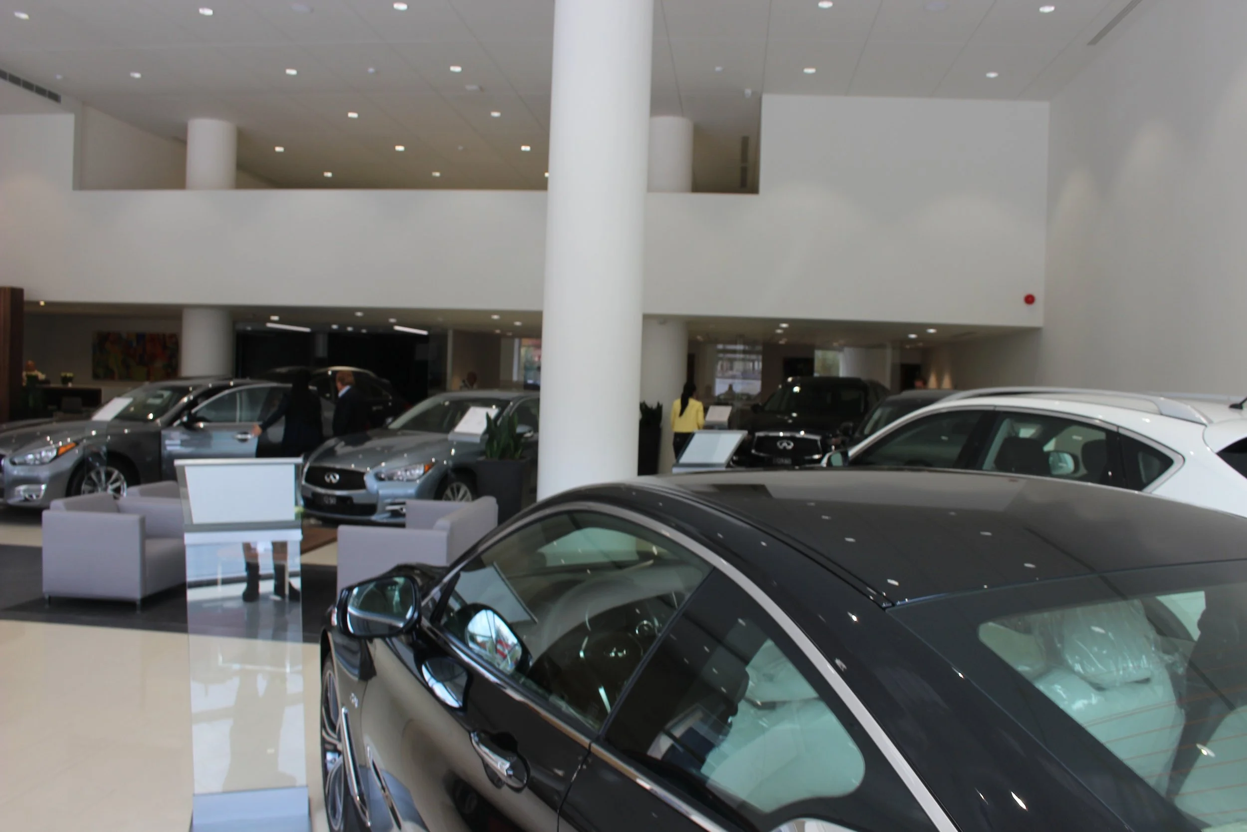 Several cars displayed inside a modern car dealership showroom with white walls and ceiling, and a few people inspecting the vehicles.