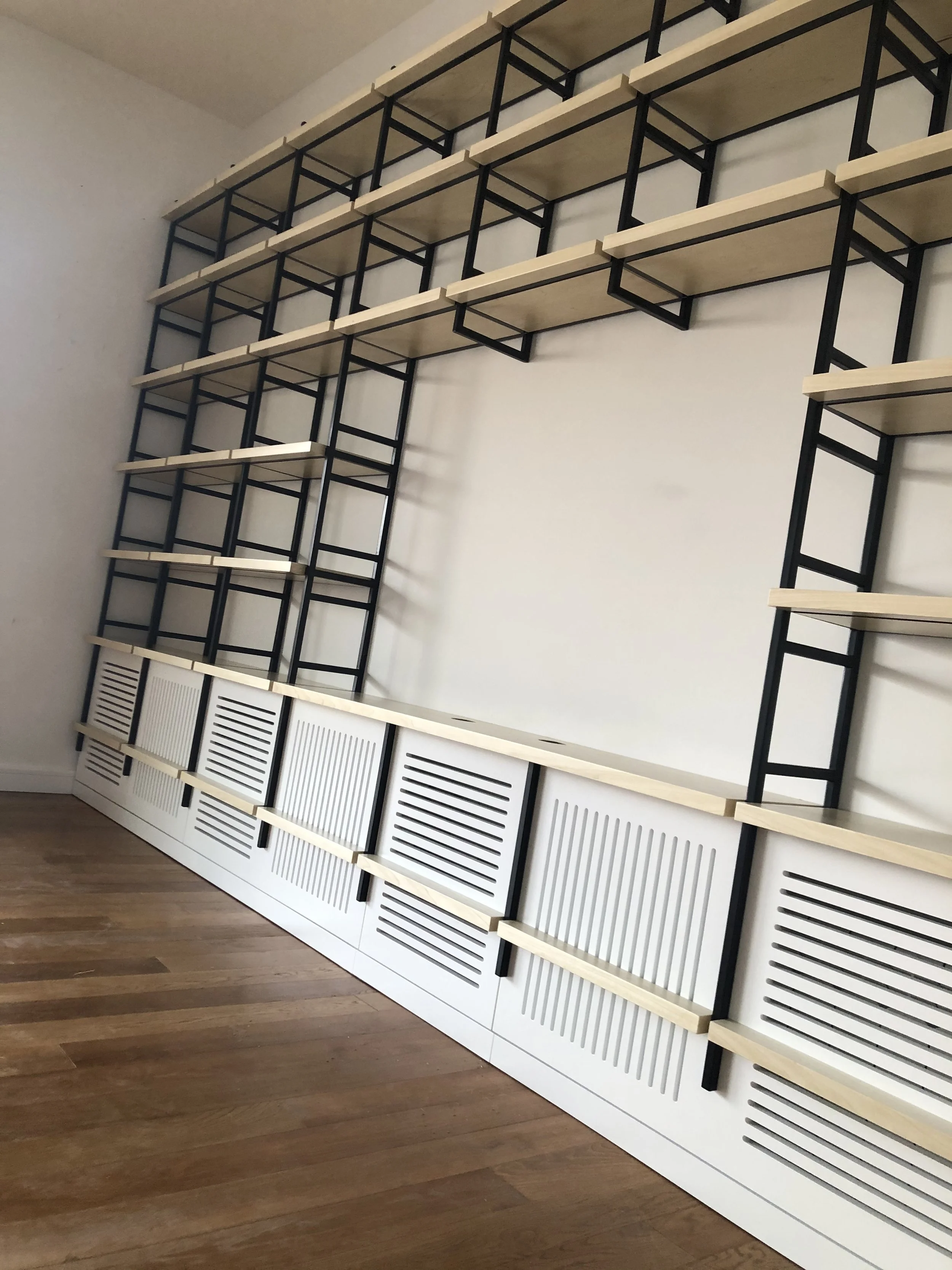A large wall-mounted shelving unit with multiple beige shelves supported by black metal frames and white louvered cabinet doors at the bottom, situated against a plain wall in a room with wooden flooring.