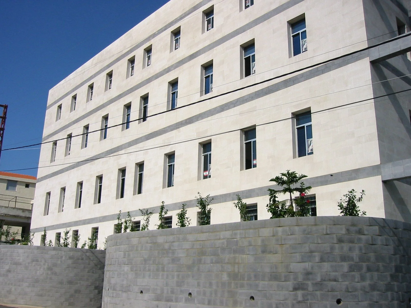 Modern multi-story building with white exterior walls, multiple rectangular windows, and a lower stone wall with small trees and plants.