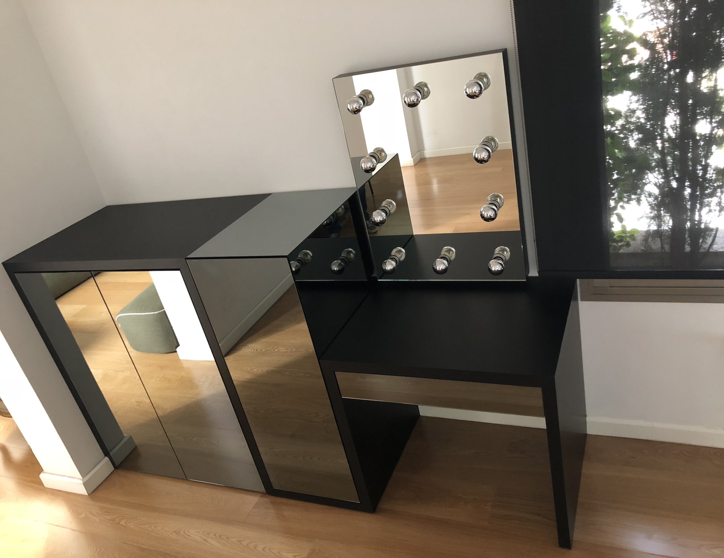 Black vanity with mirror and round metallic lamps, beside a window with trees outside, in a room with wood flooring.