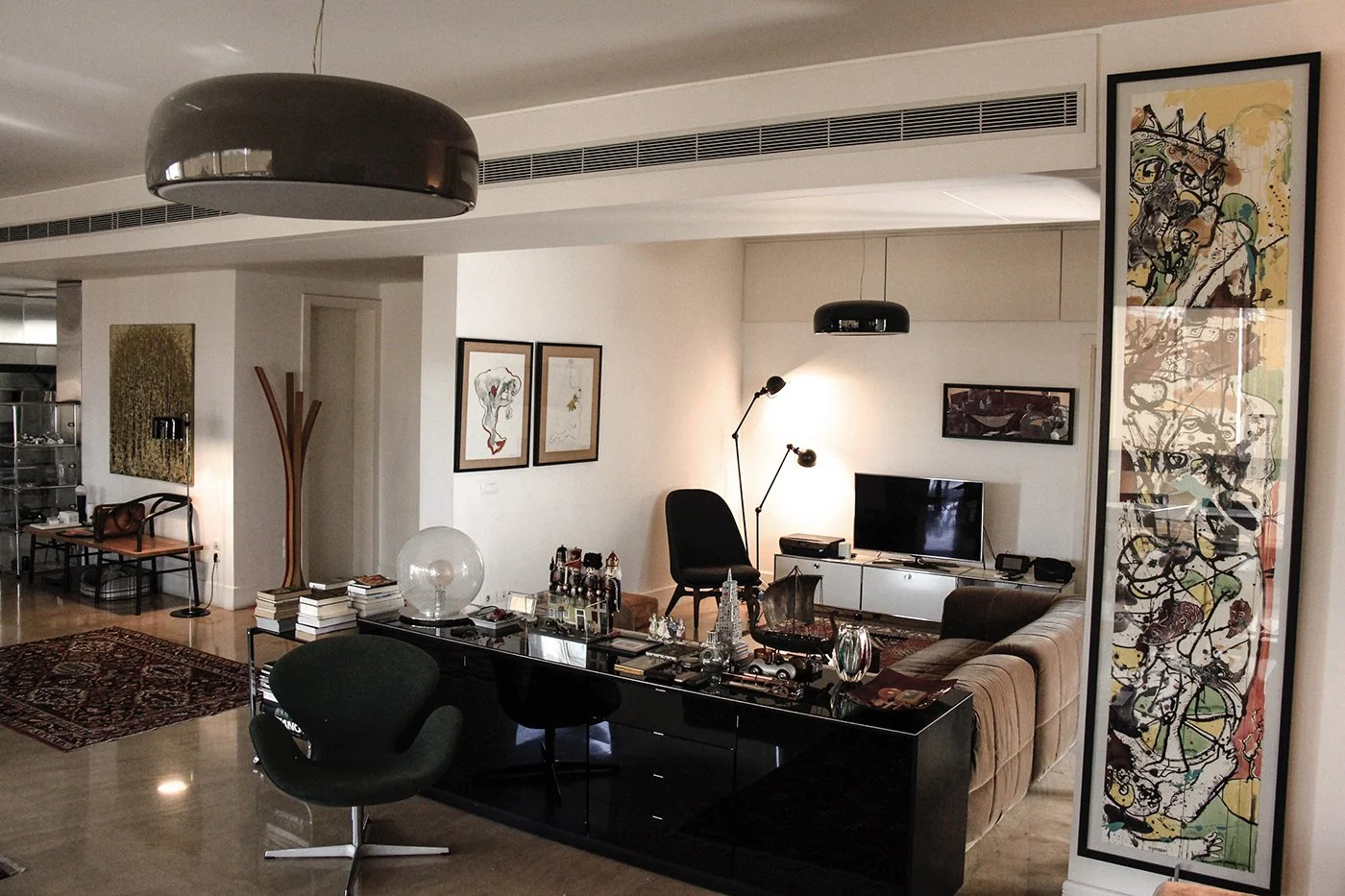 Interior of a modern living room with a brown sofa, black armchair, white TV stand, wall art, decorative objects on a black console, and contemporary lighting fixtures.