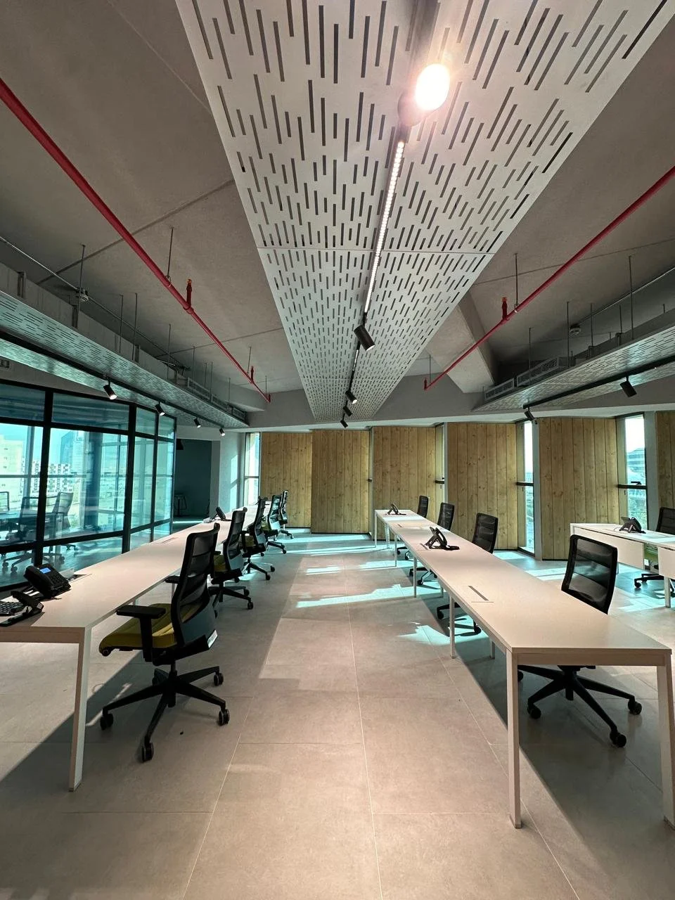 Empty modern office meeting room with white desks, black office chairs, telephones, large windows letting in natural light, and a decorative ceiling with linear patterns.