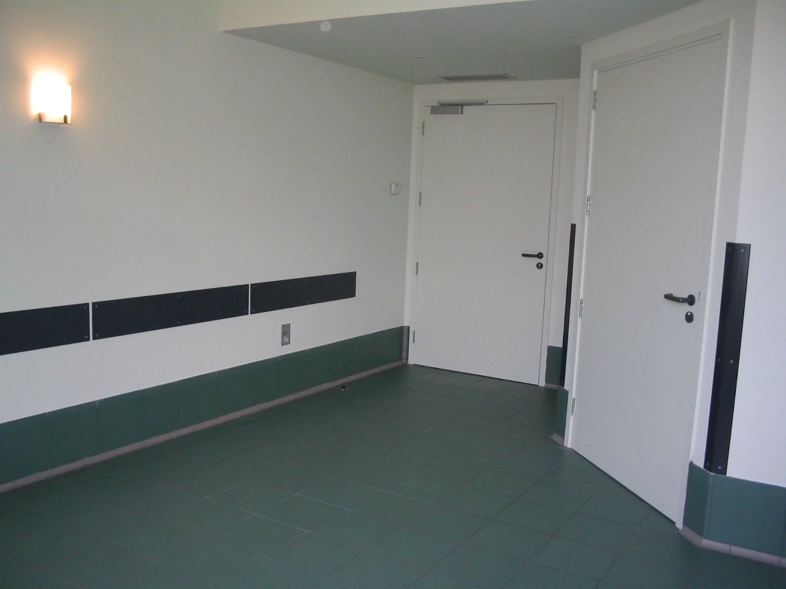 Interior of a hallway or corridor with white walls, green tiled floor, two white doors, one with an electronic lock, a wall-mounted light fixture, and black horizontal panels along the wall.